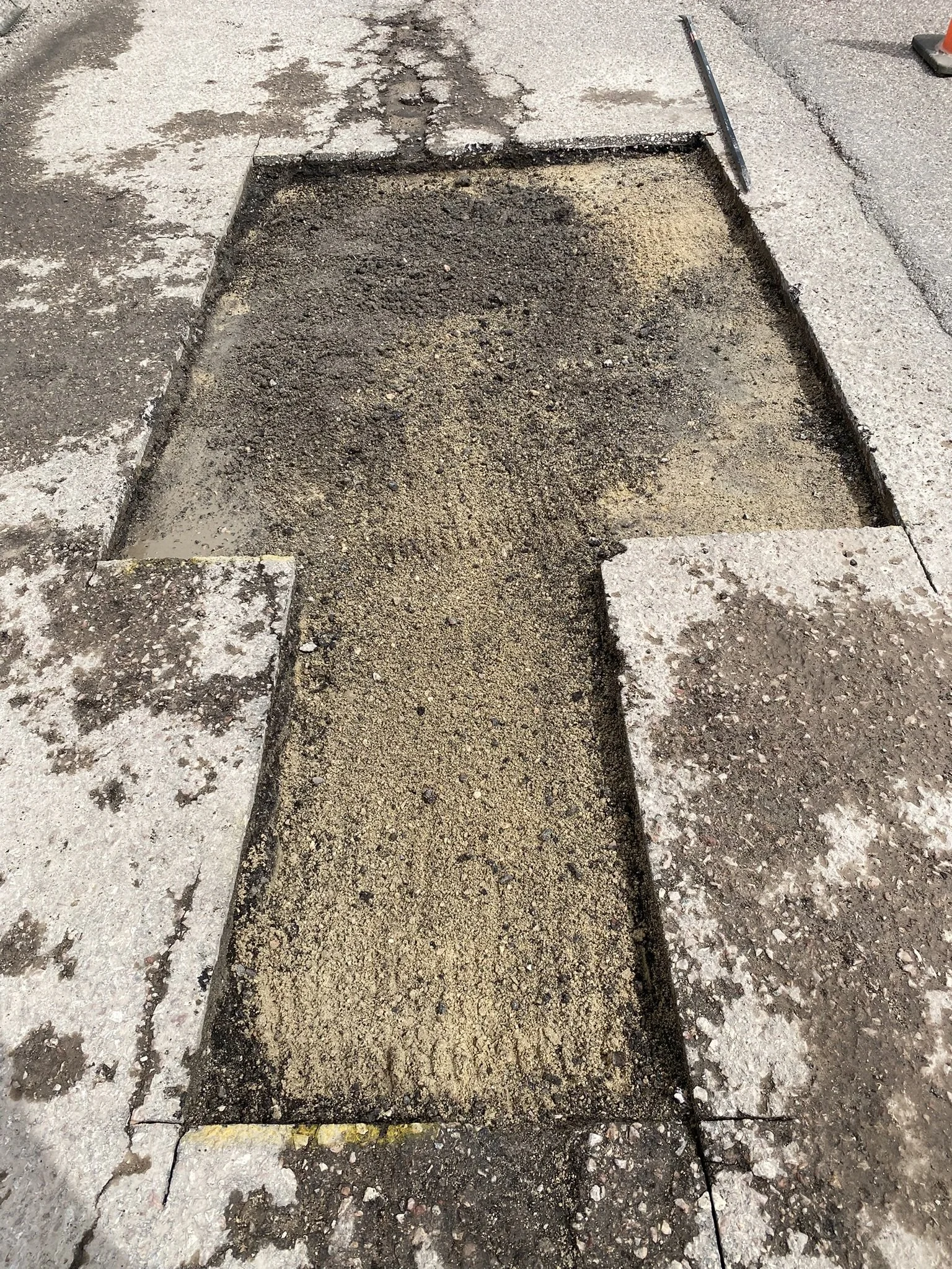 A section of a sidewalk removed for repair, exposing the underlying dirt and gravel, with a straight edge tool placed nearby, and surrounding concrete and asphalt surfaces.