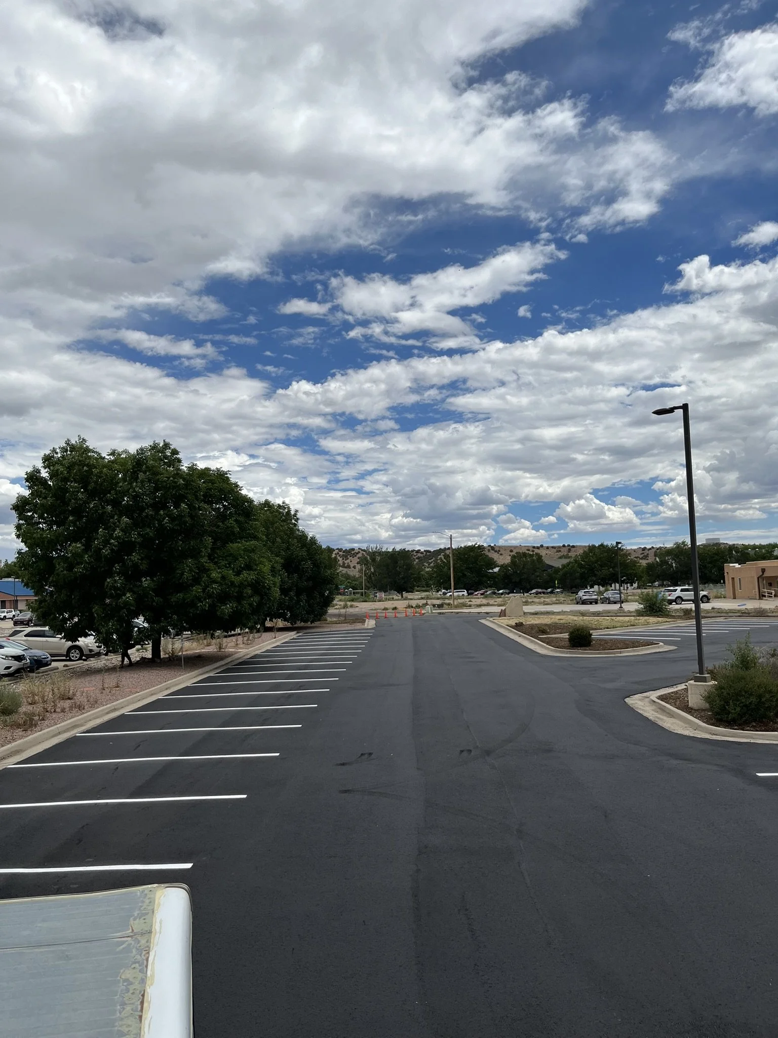 Empty parking lot with white lines, green trees, a streetlamp, and a partly cloudy sky with patches of blue.