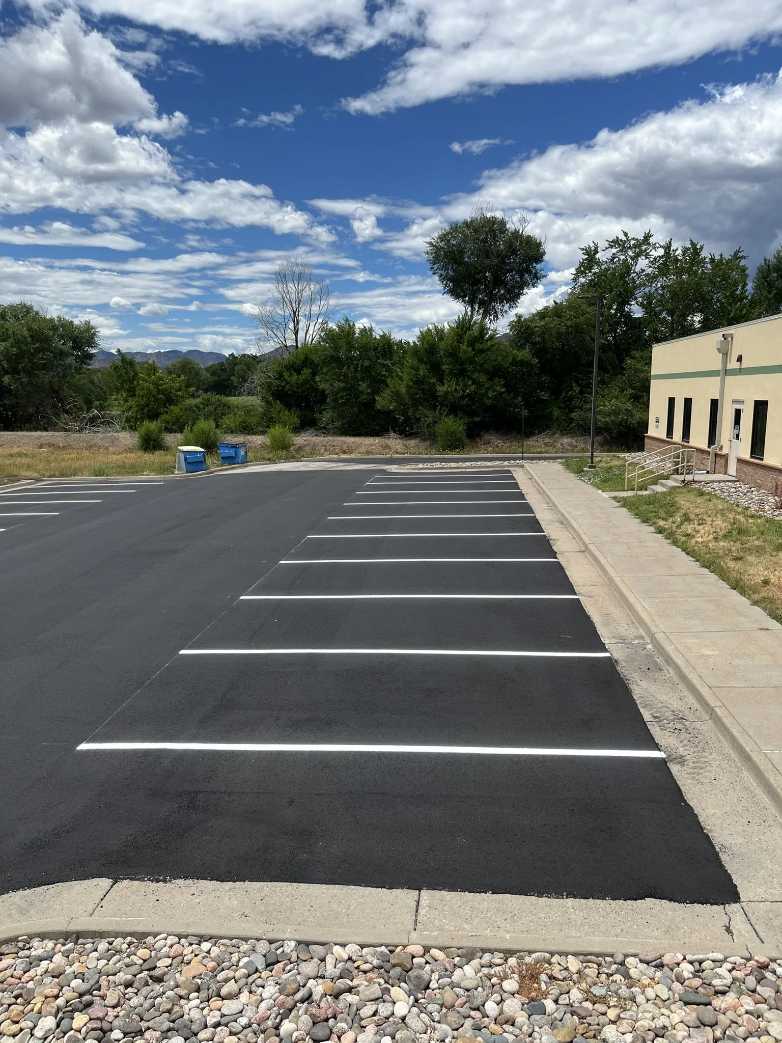 Empty parking lot with freshly painted white parking space lines, next to a sidewalk with a building on the right, trees and blue sky with clouds in the background.