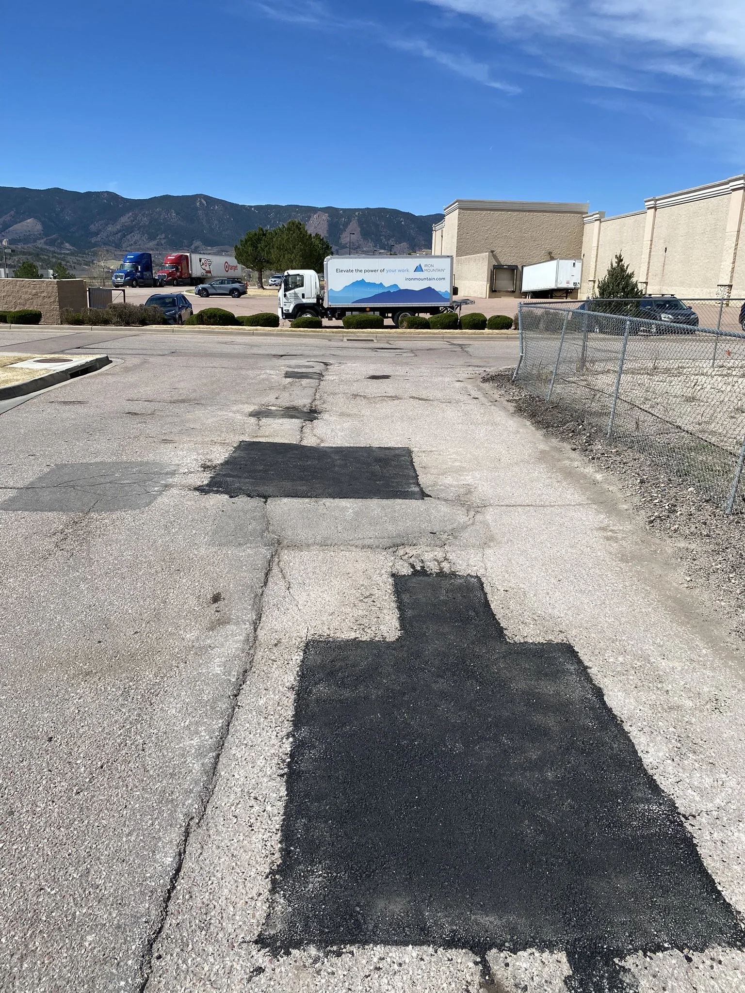 A parking lot with existing cracks and freshly repaired asphalt patches, chain-link fences, parked cars, and a building with trucks in the background, against a backdrop of mountains and a bright blue sky.