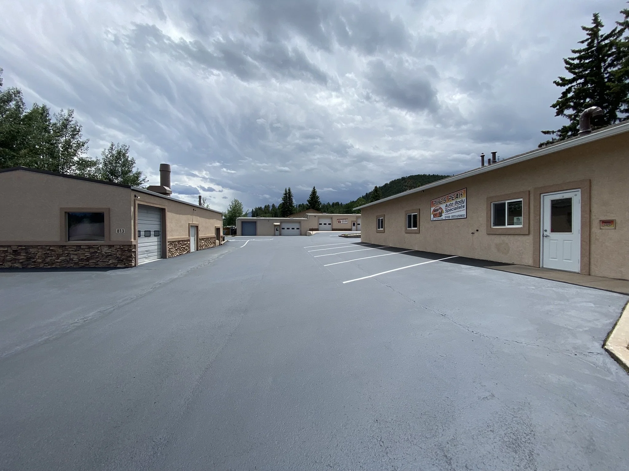 A commercial building with a sign for Pikes Peak Auto Body Specialists, situated in a parking lot with empty spaces, surrounded by trees and mountains under a cloudy sky.