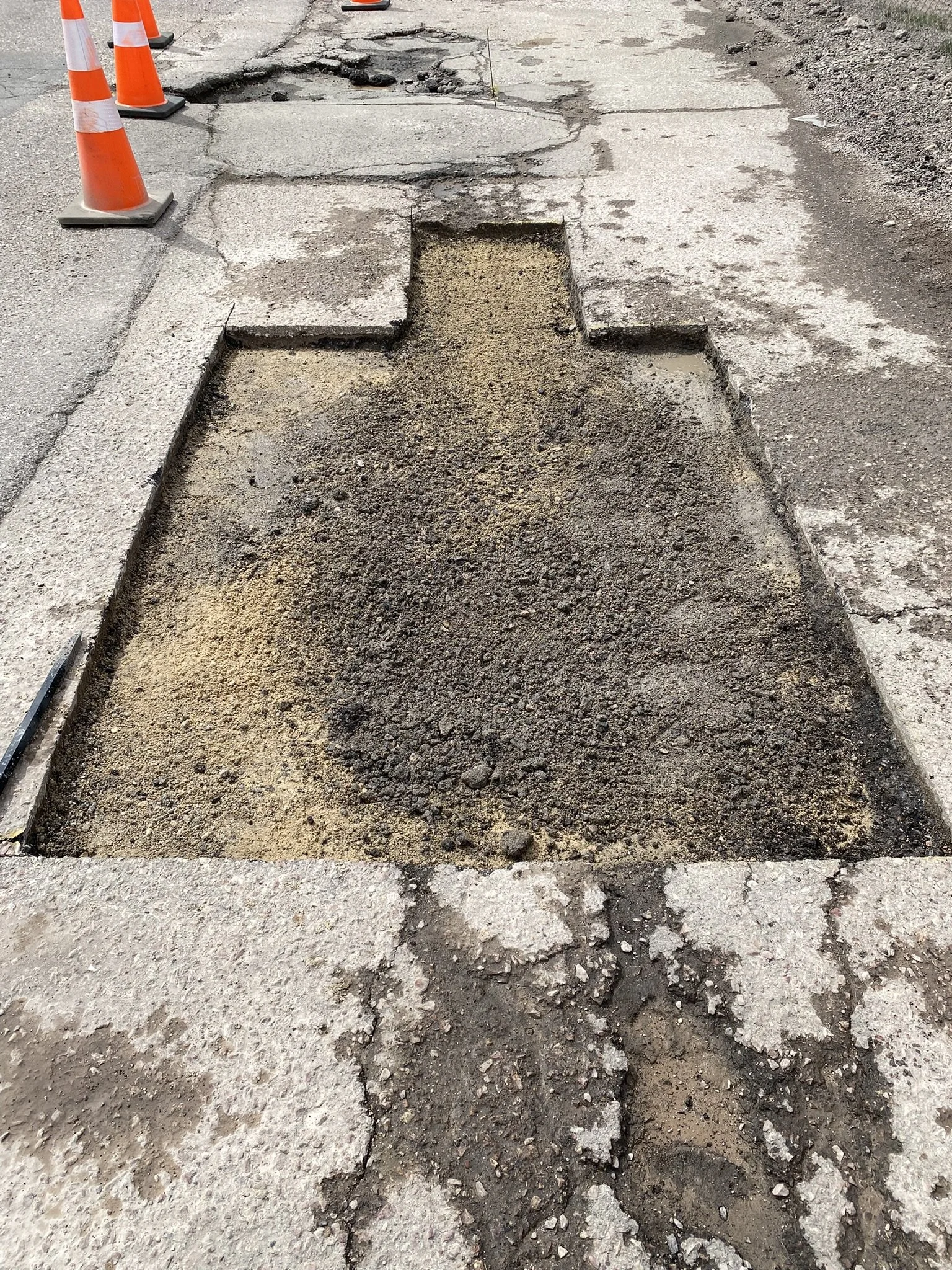 Part of a sidewalk with a rectangular section dug out, exposing dirt and gravel underneath, with construction cones on the side.