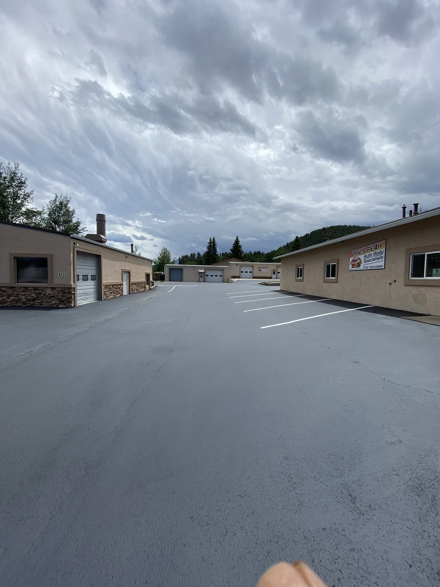 A parking lot with several small beige buildings under a cloudy sky, with a mountain in the background and parked cars.