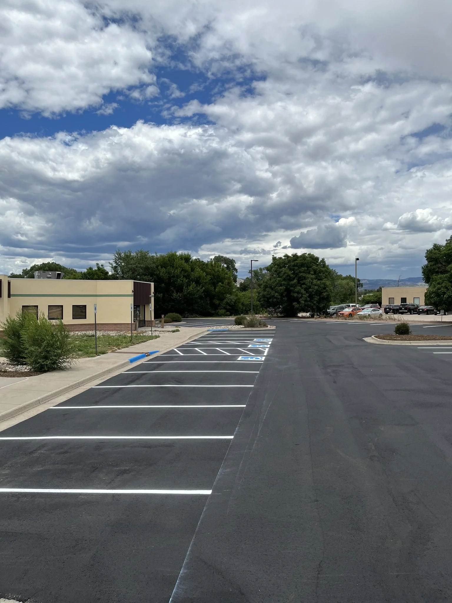 Empty parking lot with some parking spaces marked, a small building, trees, and a cloudy sky with patches of blue.