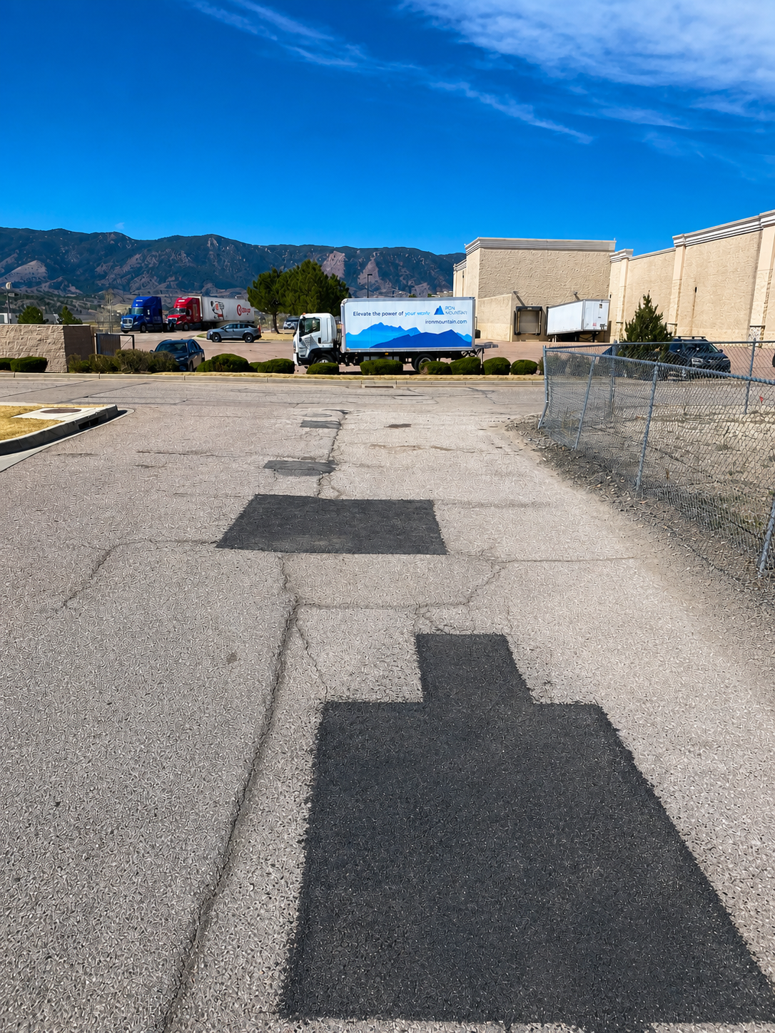Street view with black patches on the pavement, chain-link fence on the right, vehicles including trucks and cars parked in the background, and mountains under a clear blue sky.