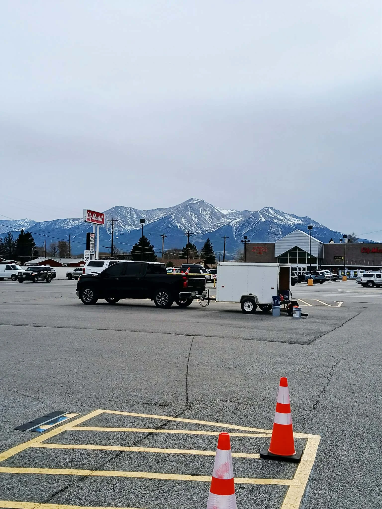 Parking lot with two orange and white traffic cones, a small trailer, and a mountain range in the background with snow-capped peaks.