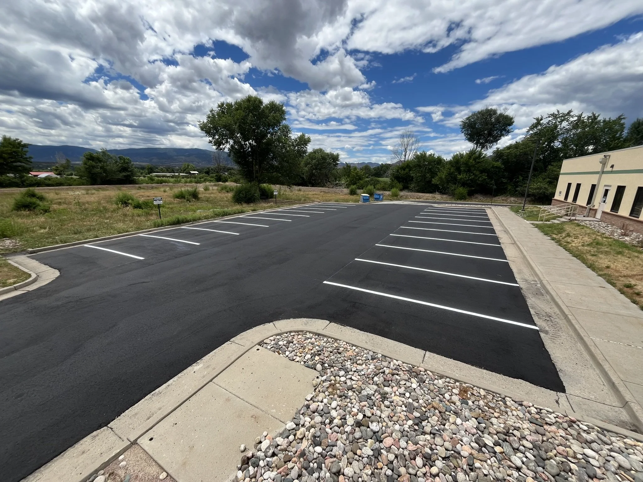 Empty parking lot with newly paved lines and landscaped curb with rocks, trees, and mountains in the background under a partly cloudy sky.