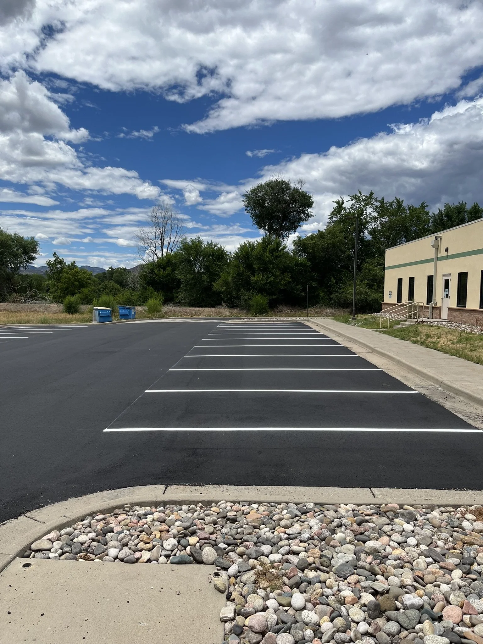 Empty parking lot with marked parking spaces, a sidewalk, rocks, trees, a building, and a partly cloudy sky.