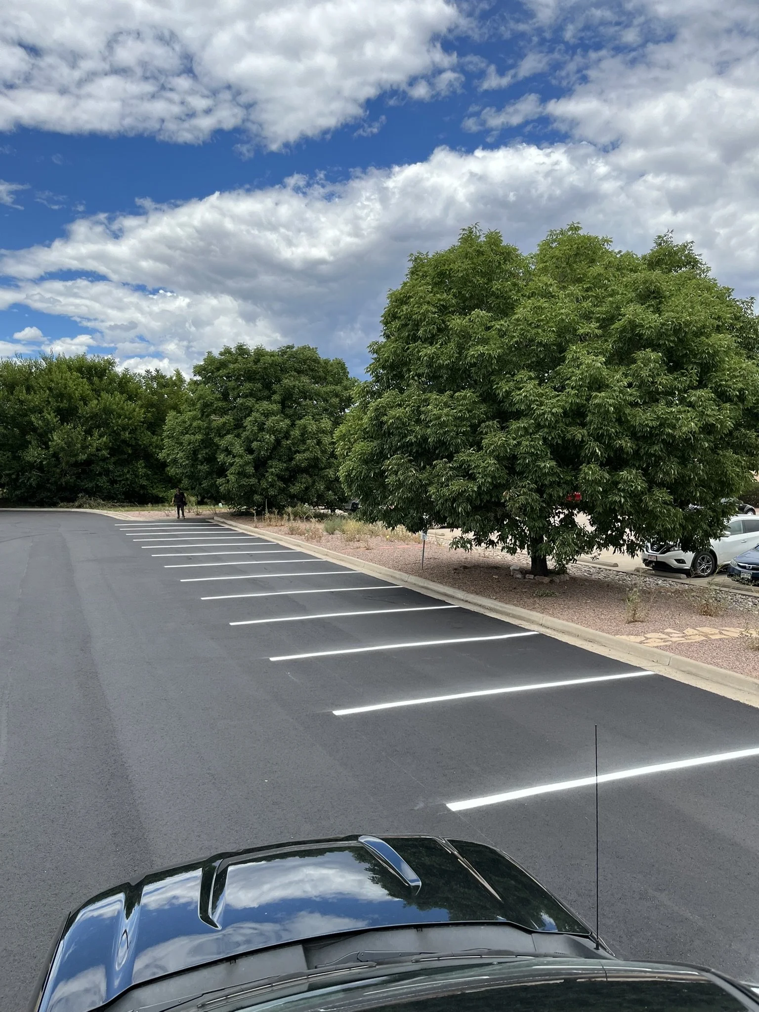 Empty parking lot with white parking space lines, green trees, a cloudy blue sky, a man standing near trees, and part of a black car at the bottom.