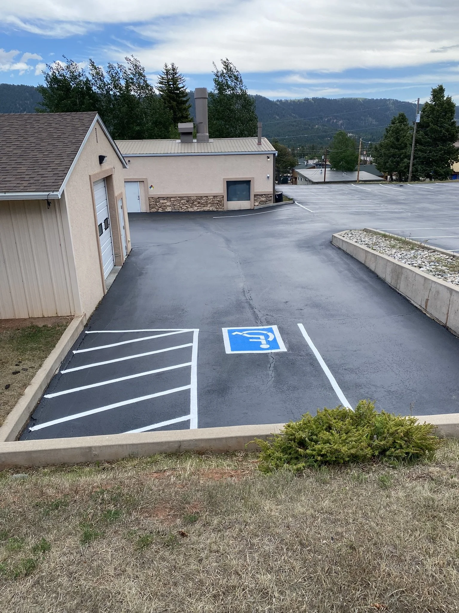 Empty parking lot with designated handicapped parking space in the foreground, building with beige walls and brown roof to the left, trees and mountains in the background under a cloudy sky.