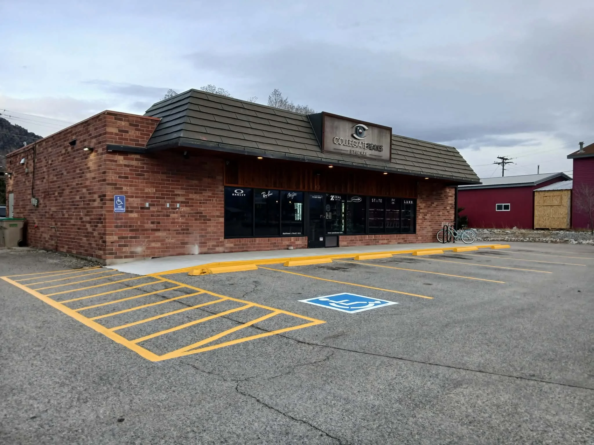 A brick building with a sign for Collegiate Eyes Eyecare on the front. There is a parking lot with a handicap space marked on the ground and two bicycles parked nearby. The sky is cloudy.