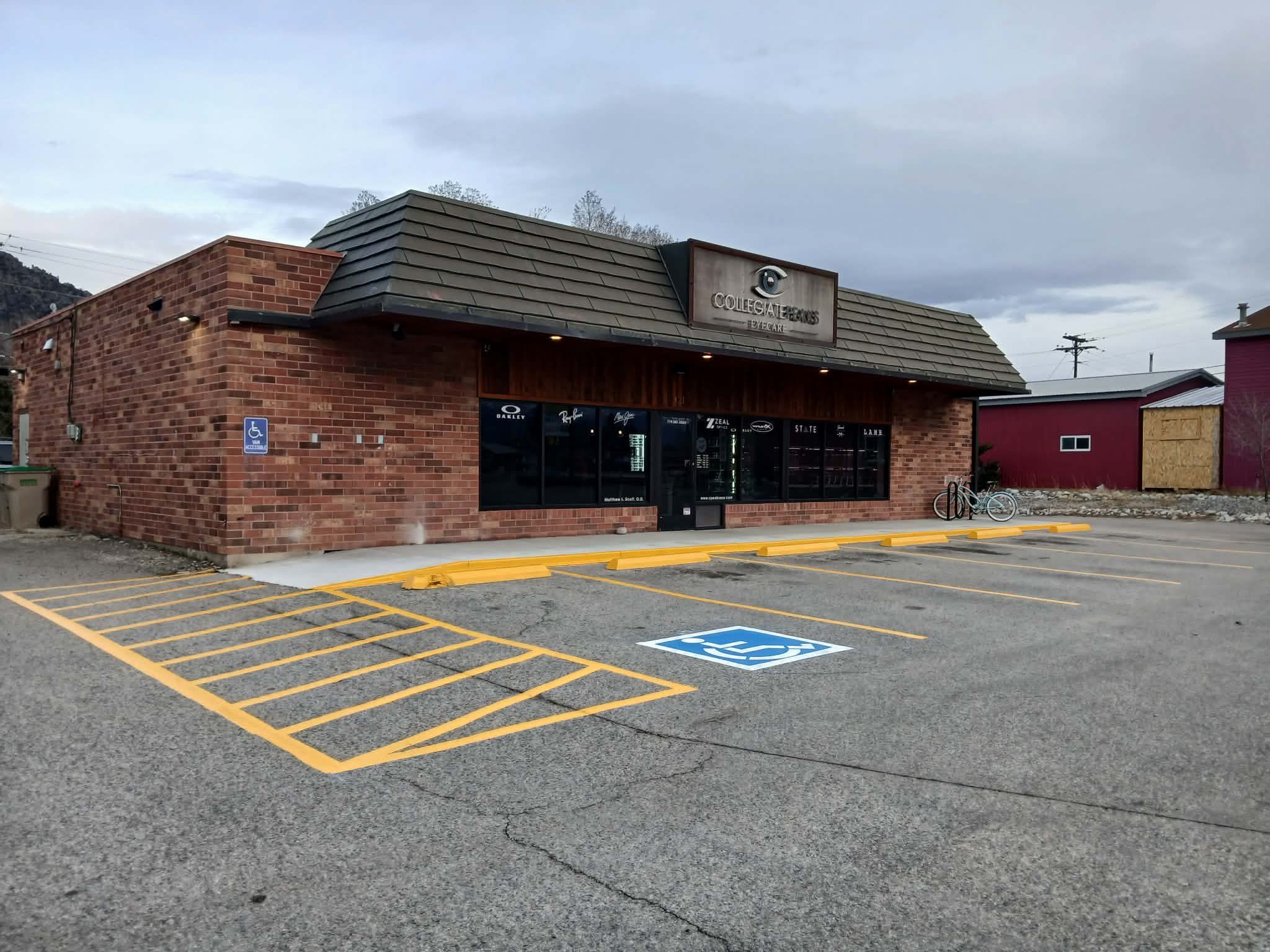 A brick building with a sign that says 'Collegiate Eyewear Eyecare' and a logo of an eye. There are parking spaces in front, including a designated handicapped parking spot. The parking lot has yellow markings and an asphalt surface, with a bicycle p