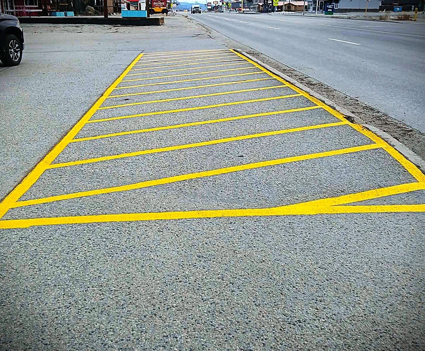 Empty parking space outlined with yellow lines on a parking lot near a city street, with other vehicles and buildings visible in the background.