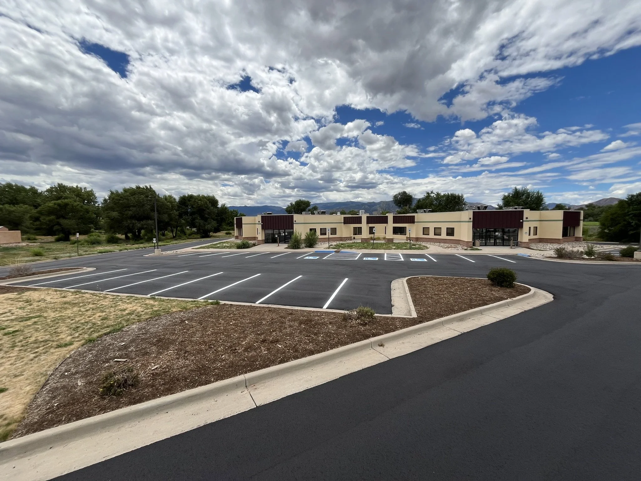 Empty parking lot in front of a modern building with mountains and trees in the background under a partially cloudy sky.