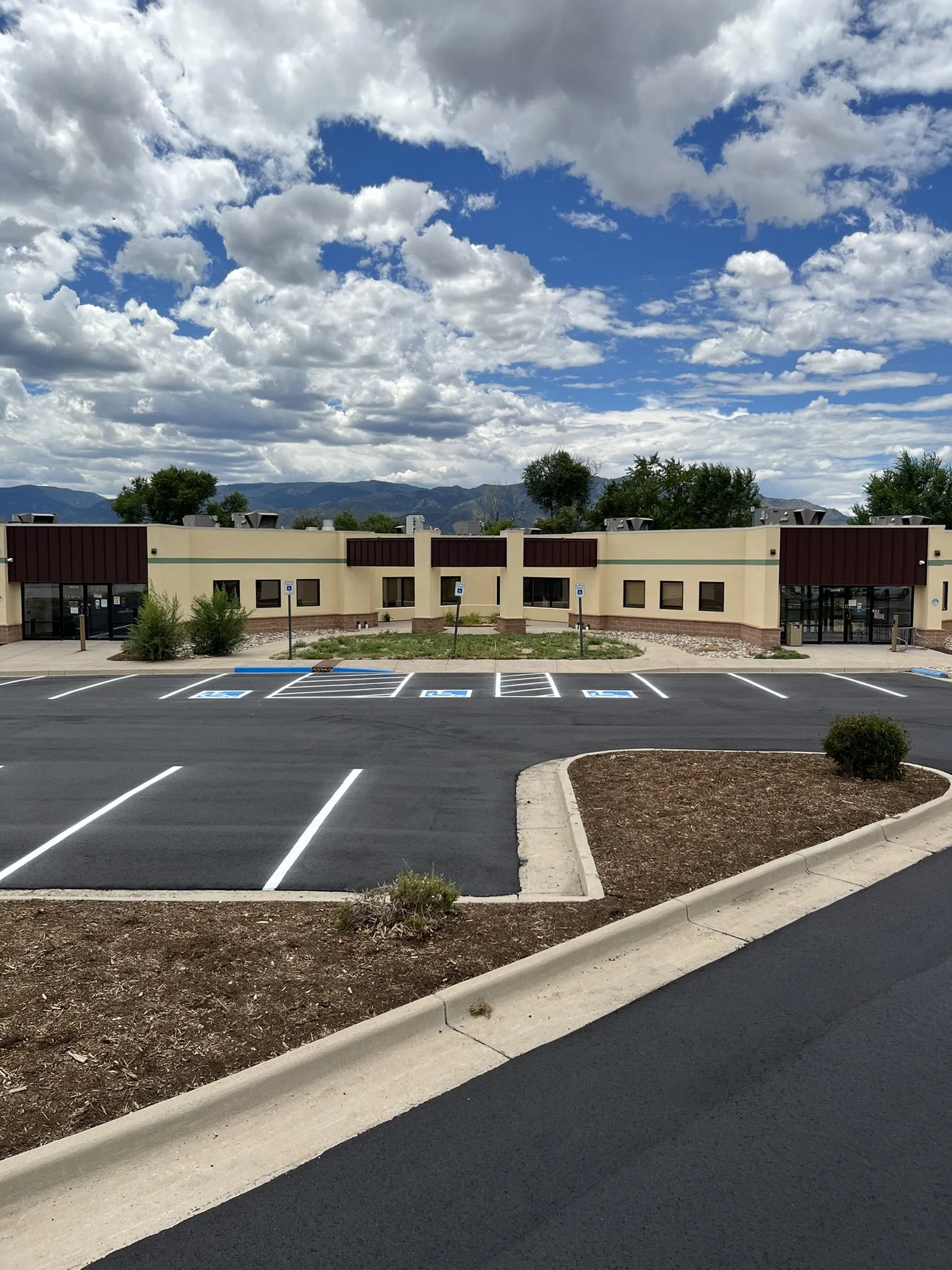 A parking lot with empty spaces, including designated handicapped parking spots, in front of a commercial building with a beige exterior and red accents. There are small bushes and a landscaped area with mulch, and trees and mountains can be seen in 