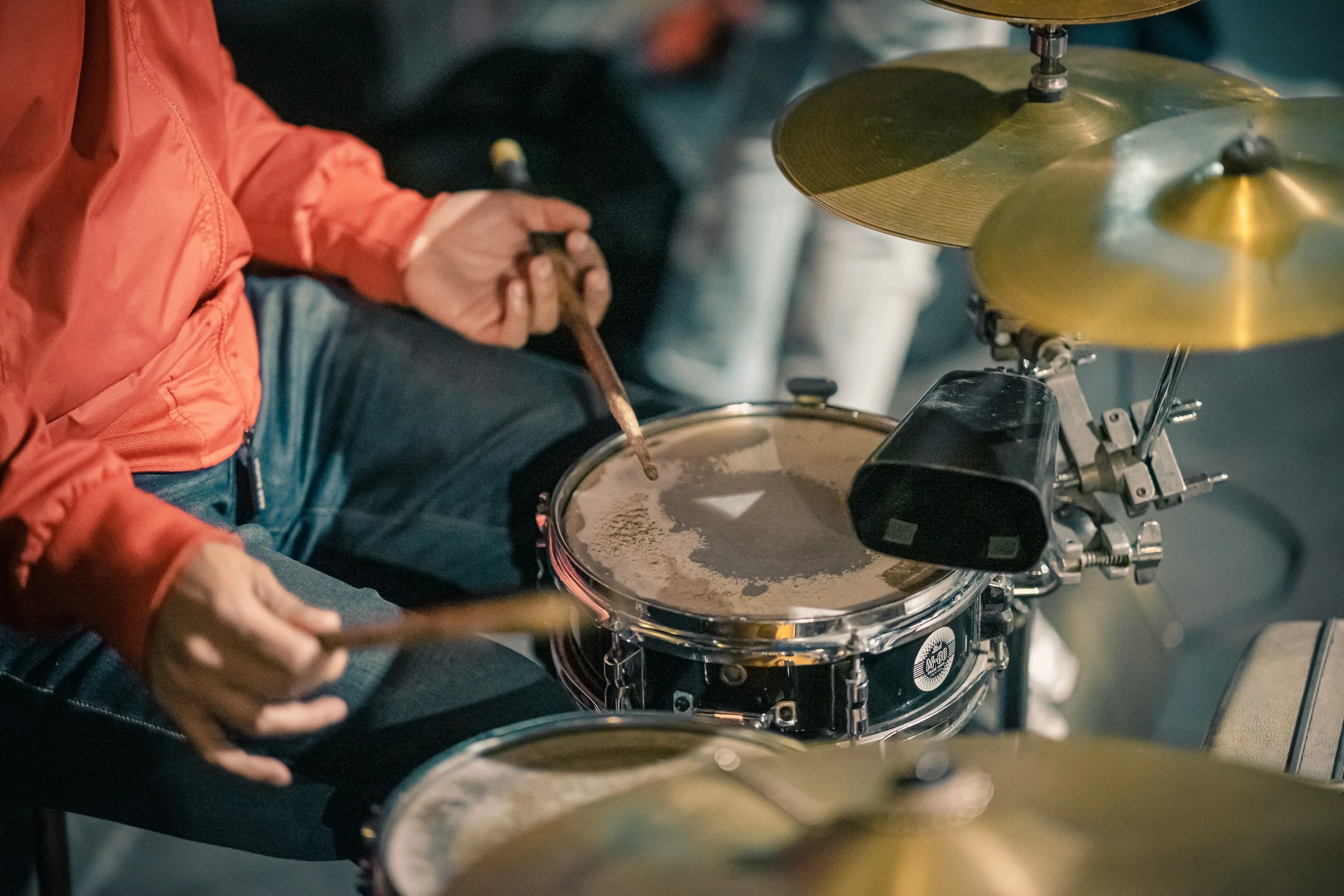 Close-up of a person wearing an orange jacket playing a drum set with brass cymbals.