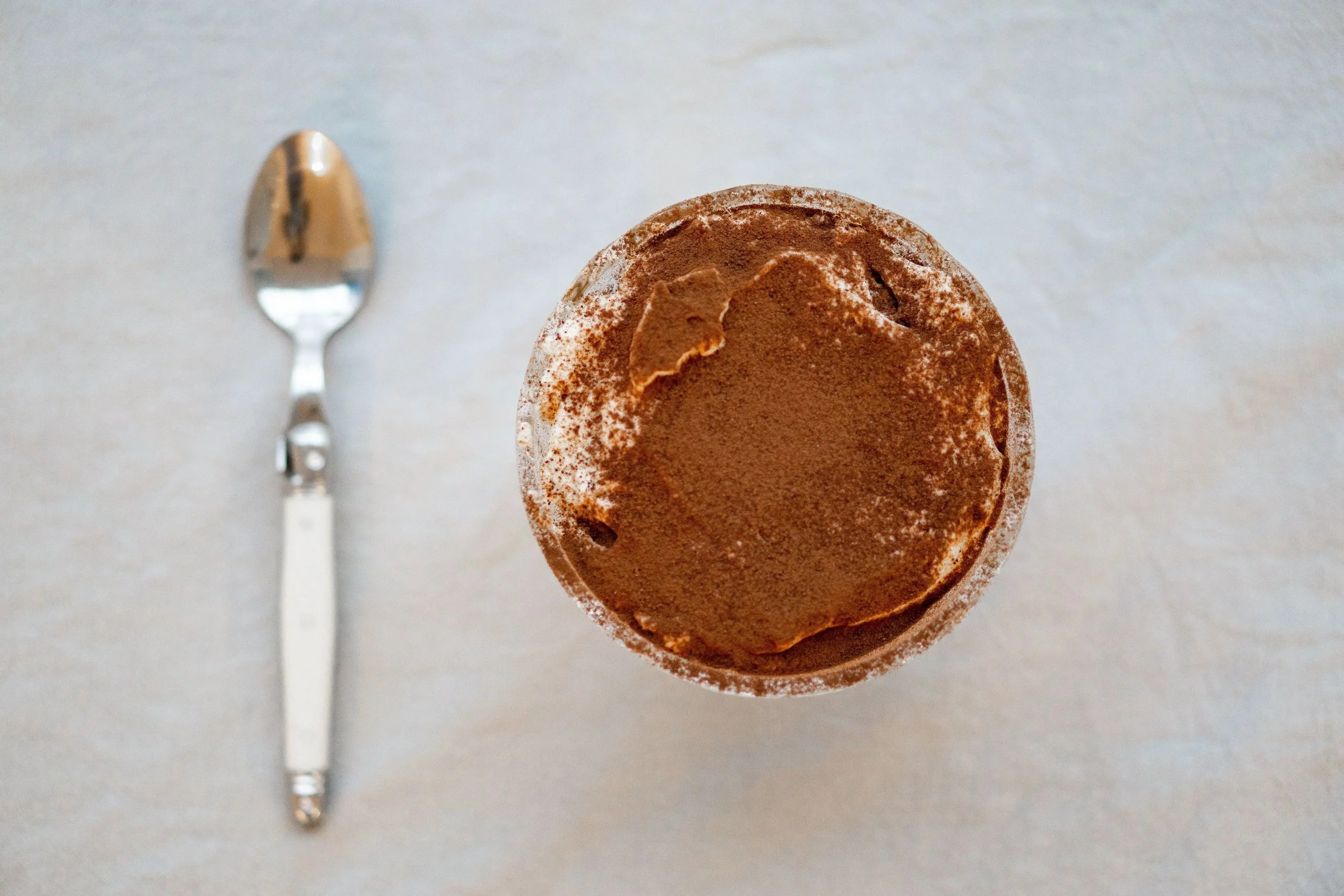 An ice cream cone with chocolate ice cream topped with cocoa powder, positioned on a white surface with a metallic spoon nearby.