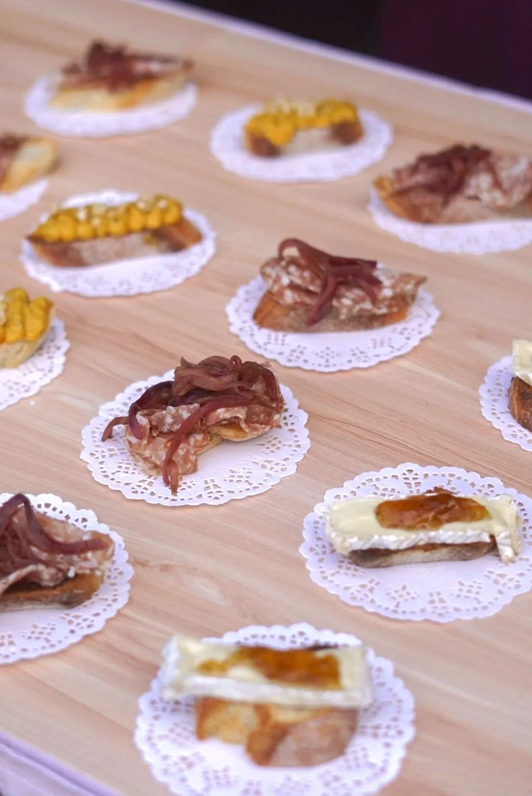 Several small appetizers on white doilies arranged on a wooden surface, including pieces with cheese, bacon, and caramelized onions.