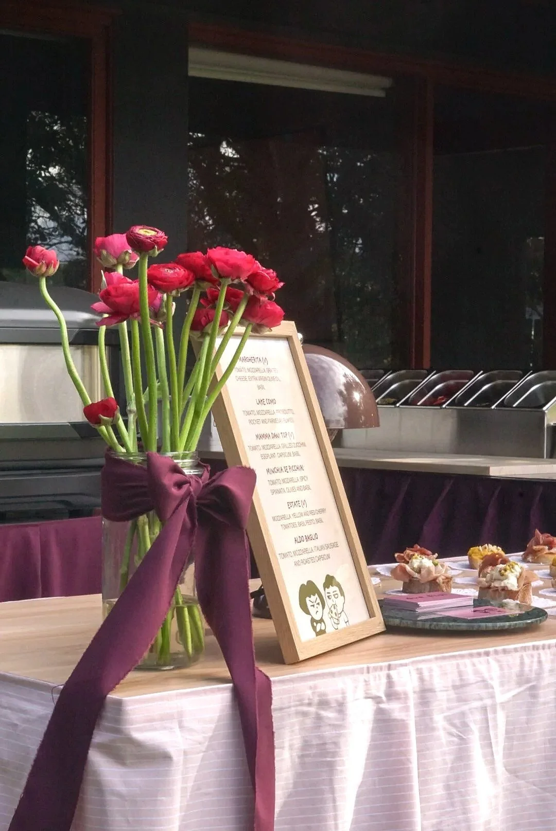 A table with a vase of pink flowers decorated with a purple ribbon, a menu on a stand, and small desserts on a tray, set against a background of a buffet or serving area and window reflections.