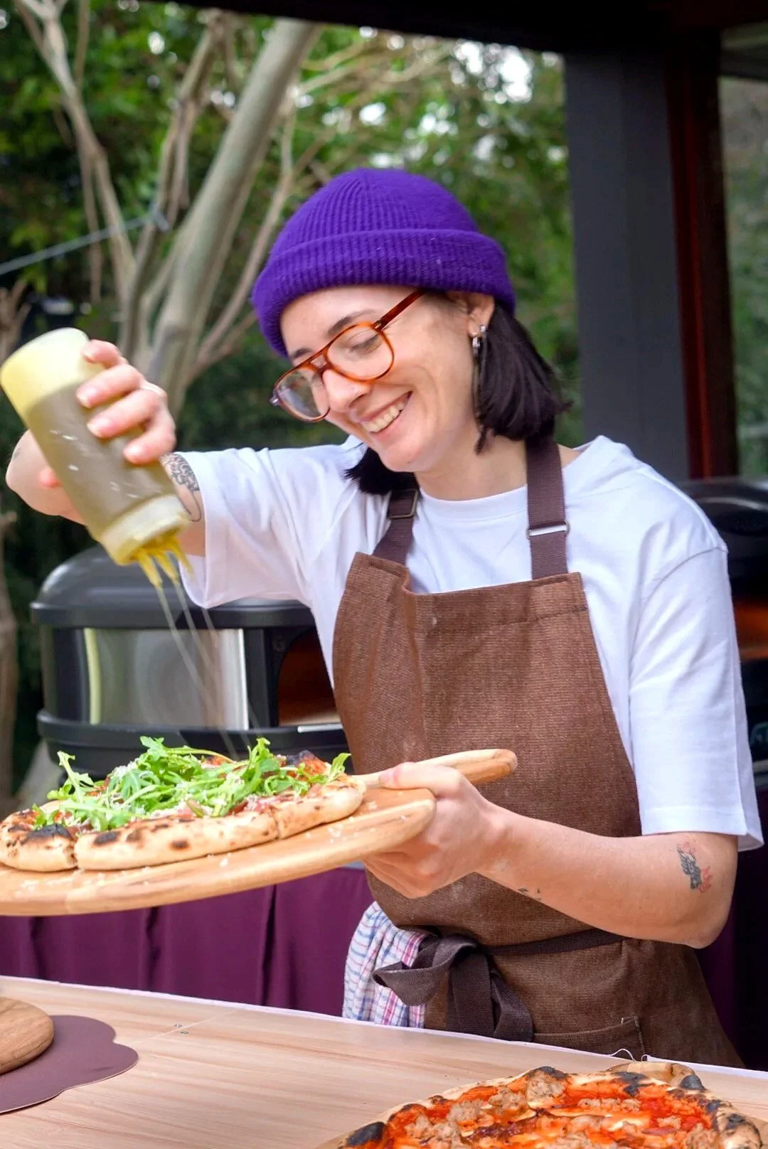 A woman with glasses and a purple beanie preparing to serve salad and pizza outdoors.