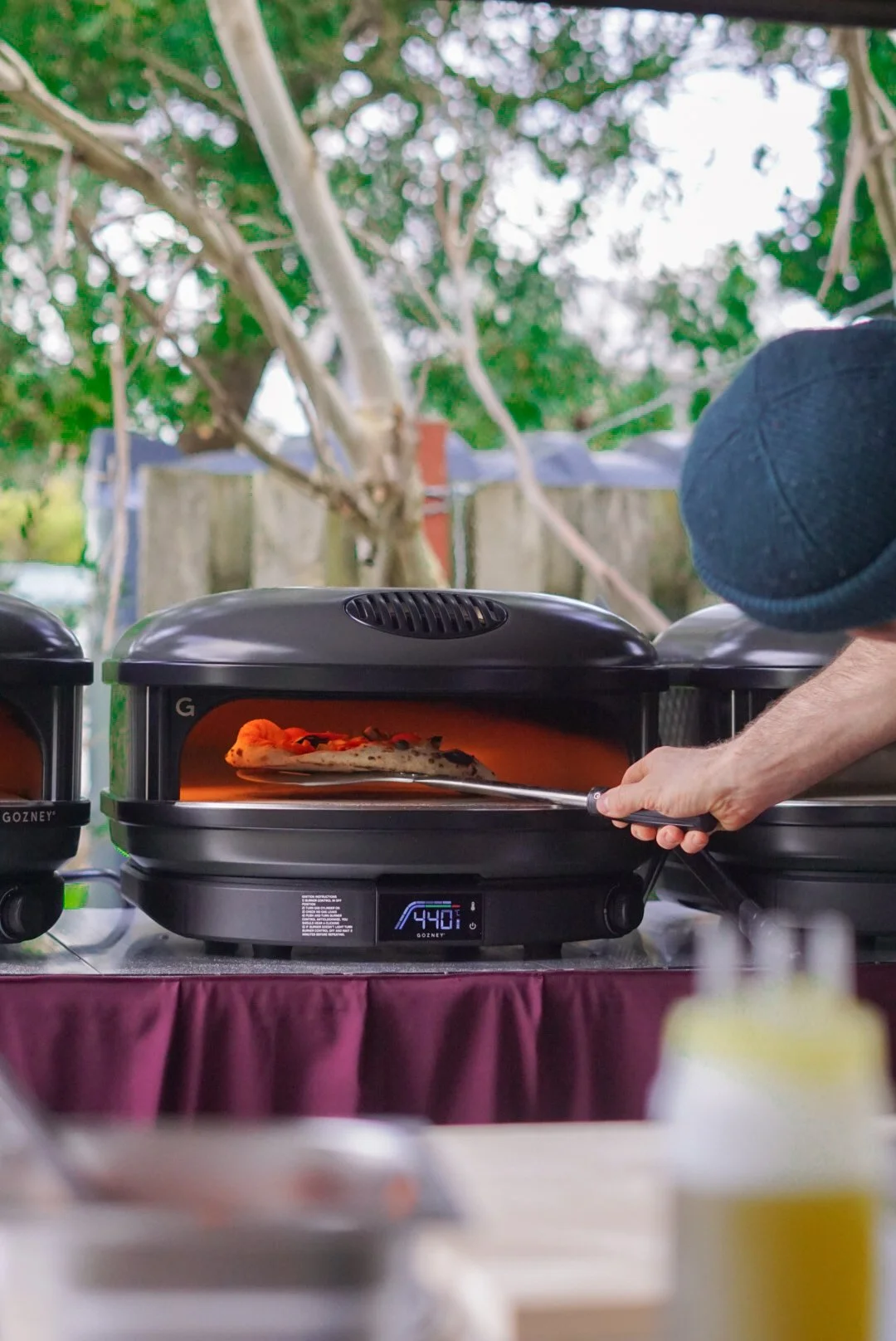 A person is removing a pizza from a black electric pizza oven outdoors, with trees and a fence in the background. The oven displays 440°F on its digital screen.