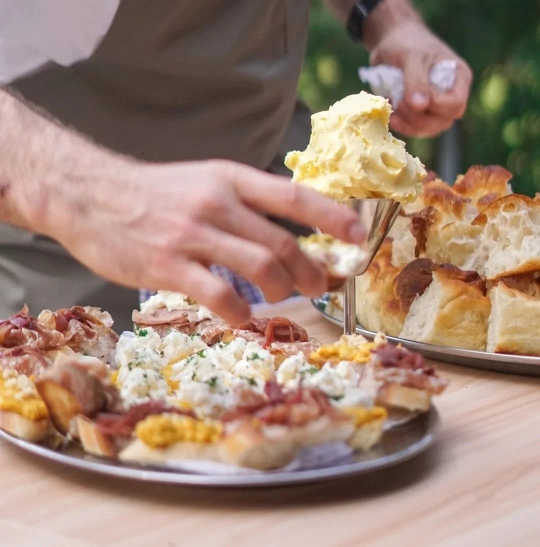 A person with light skin serving mashed potatoes onto a plate of assorted appetizers, with a tray of bread on the side, outdoors with blurred green foliage in the background.