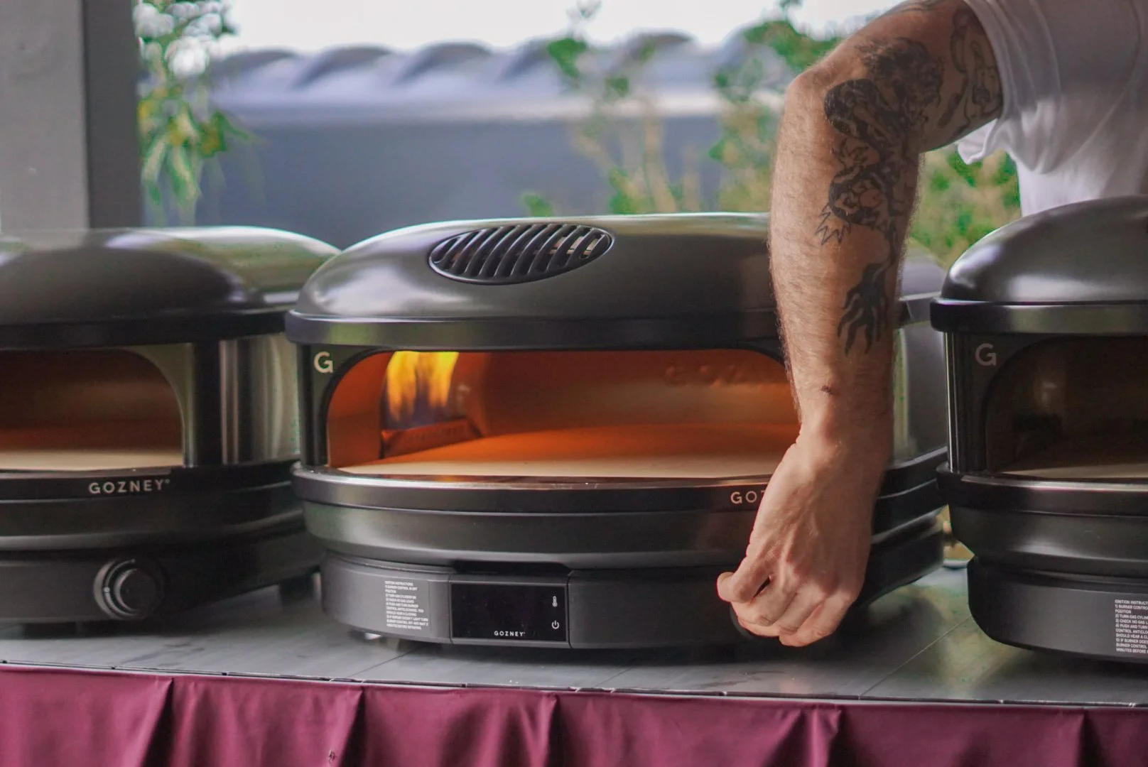 Person with tattoos using a portable pizza oven to cook a pizza, with other ovens on either side, on a table covered with a red cloth.
