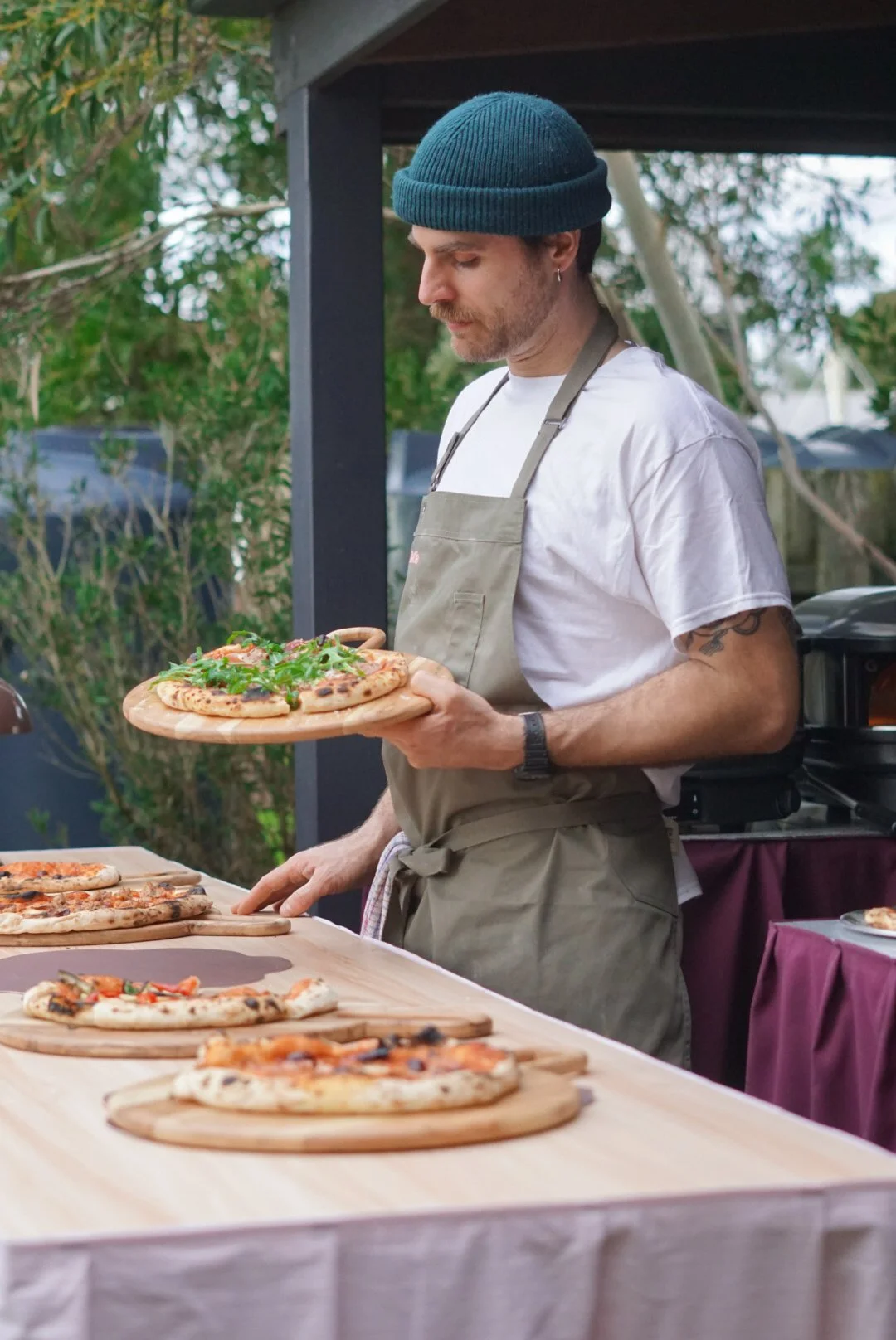 A man wearing a dark blue beanie, white t-shirt, and beige apron holds a pizza with arugula and other toppings on a wooden serving tray at an outdoor setting with trees in the background.