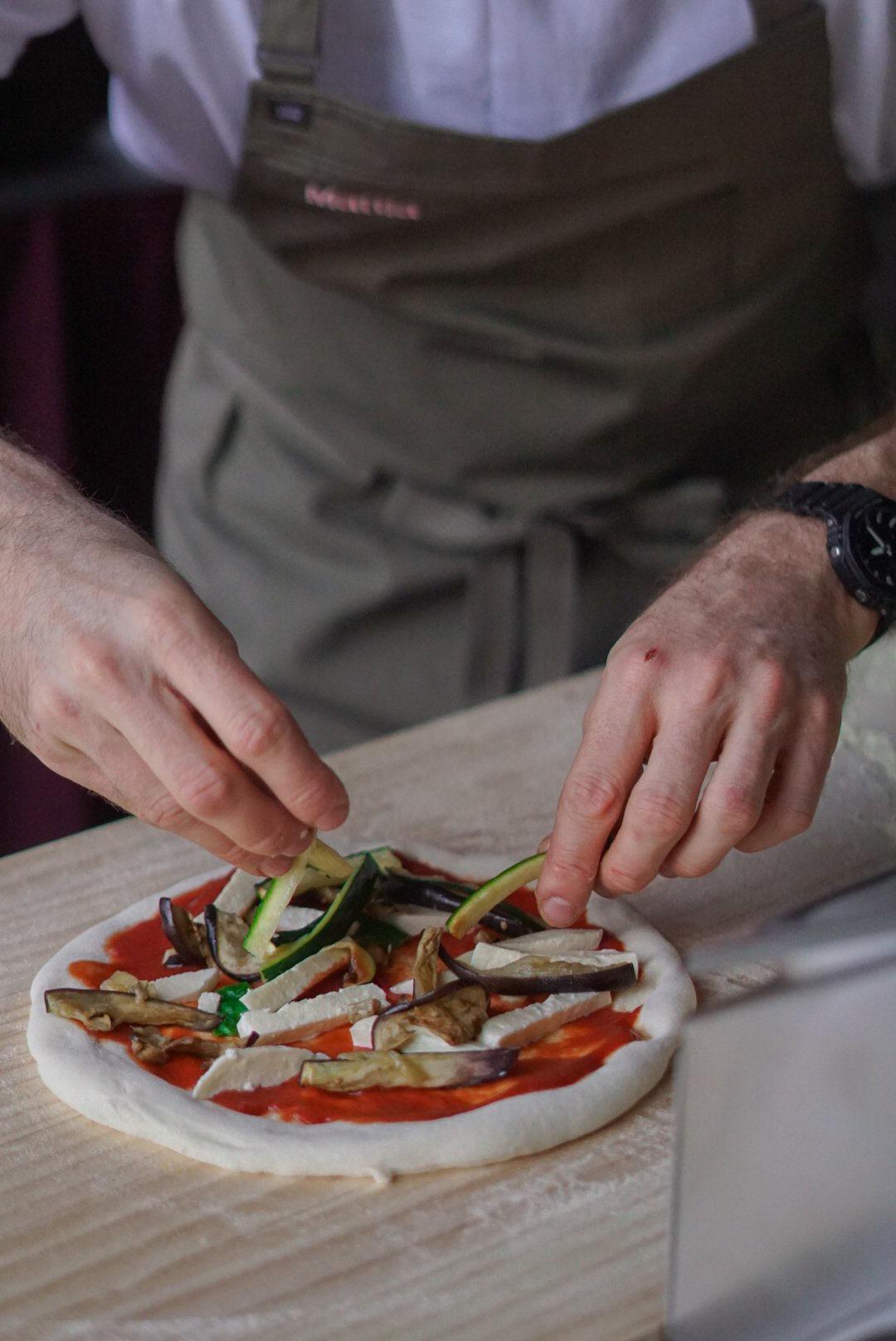 Close-up of a person preparing a pizza with vegetables on a wooden surface.