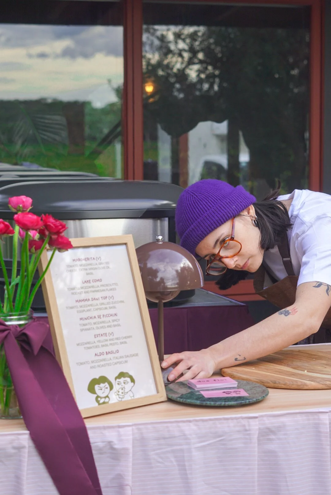 A woman with dark hair, glasses, and tattoos on her arms, wearing a purple beanie and white shirt, is leaning over a counter with a menu board and pink flowers in a vase. She appears to be preparing food or working at a food stand outside.