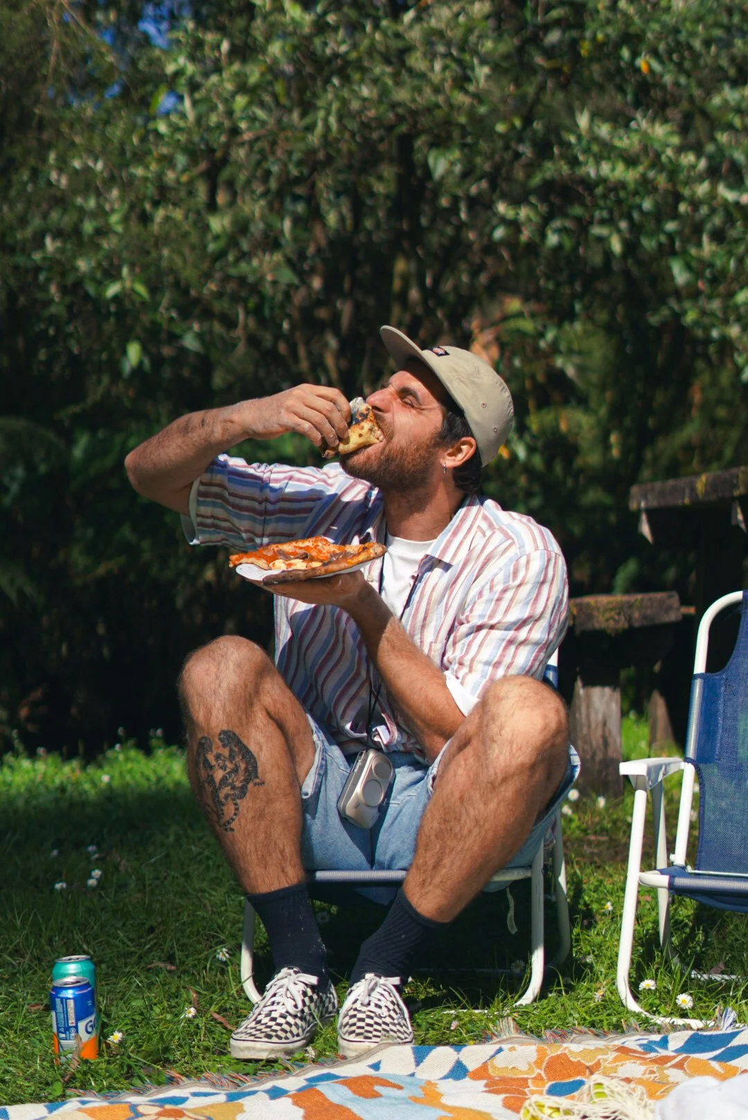 A man sitting on a camping chair outdoors, eating pizza and holding a plate with pizza slices. He is wearing a striped shirt, shorts, checkered shoes, and a cap, with a camera hanging around his neck. There are drinks on the grass and a blanket on the ground.