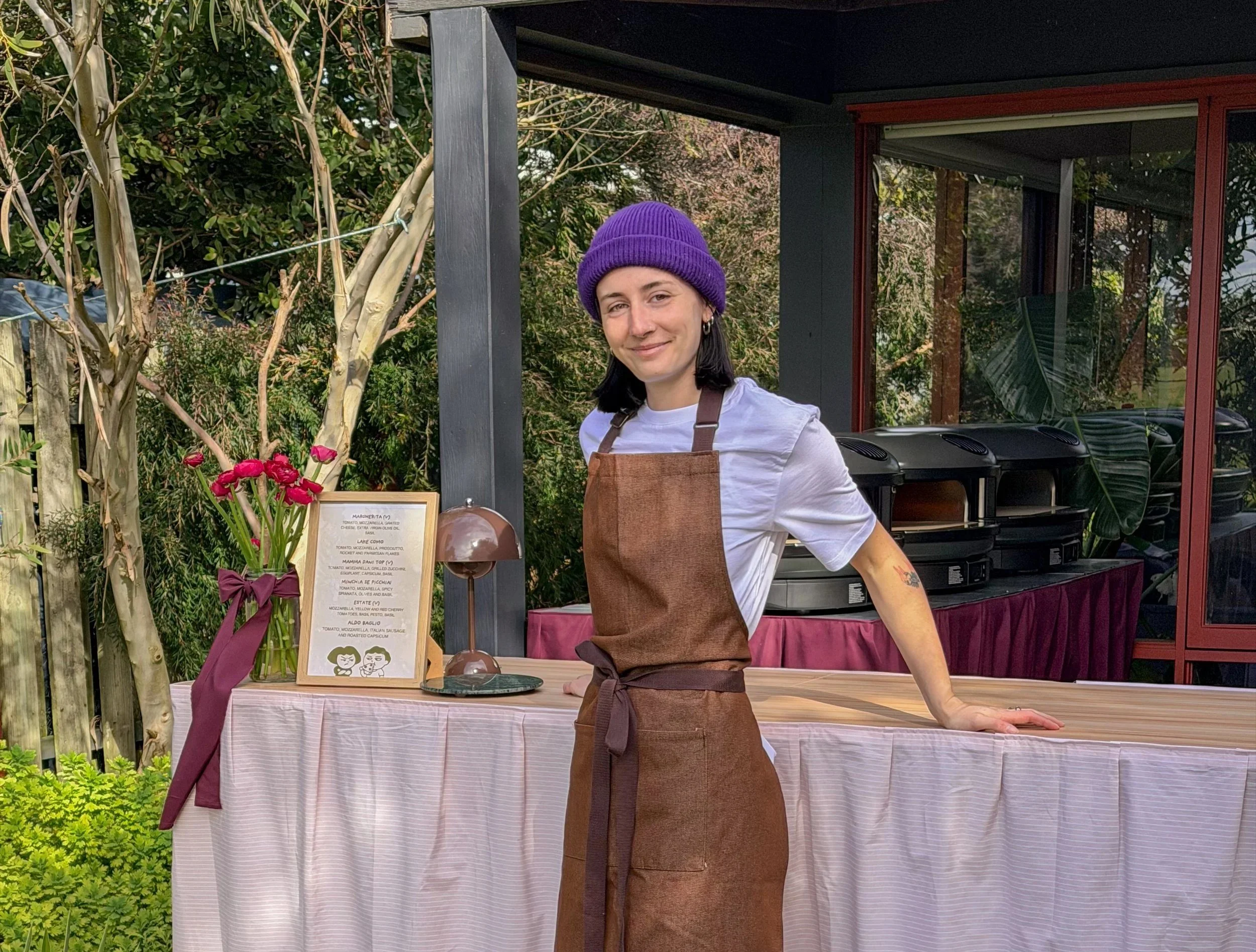 A woman wearing a purple beanie, white shirt, and brown apron standing behind a table with a pink tablecloth, outdoors near a glass building and green foliage.