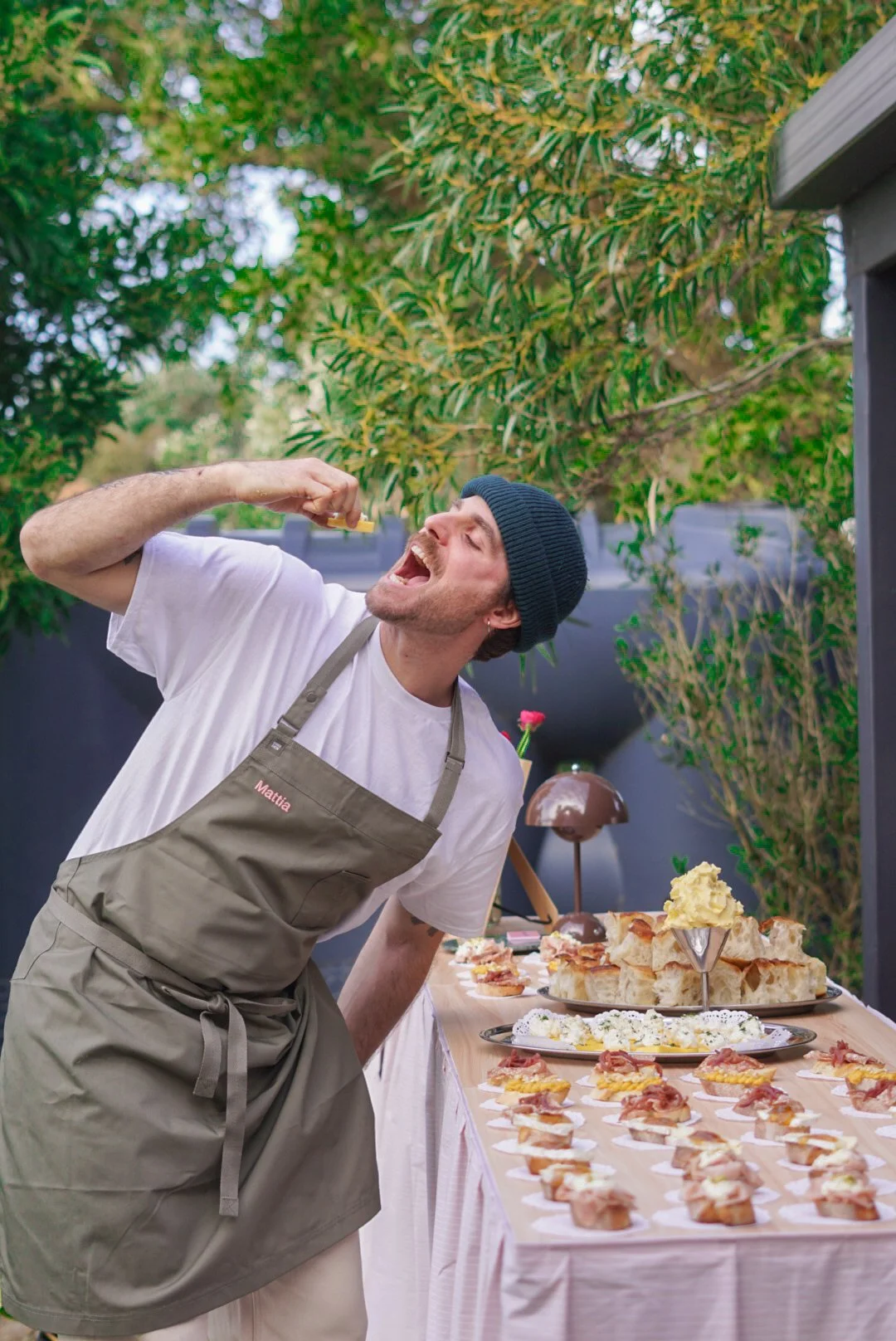 A man with a beard, wearing a blue beanie, white t-shirt, and a beige apron, is leaning back and eating a slice of food. There is a table with various appetizers, sandwiches, and a chocolate fountain in front of him, set outdoors with green foliage in the background.