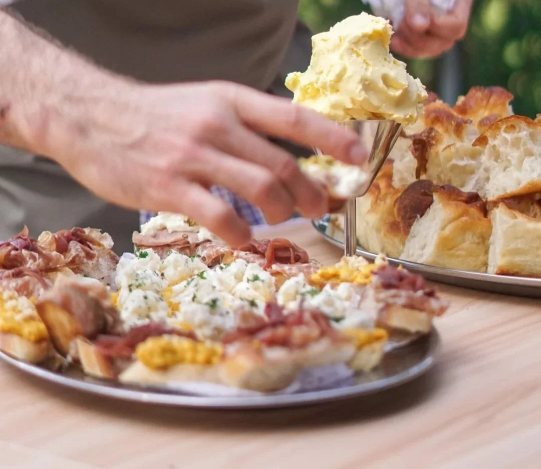 Person spreading butter on a slice of bread topped with various toppings, with more bread and toppings on a plate nearby.