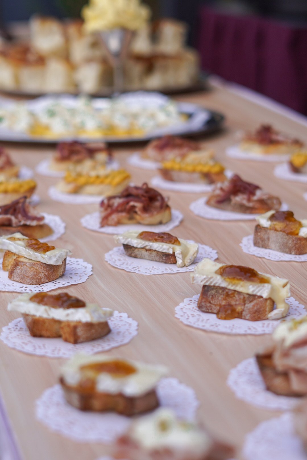 Assorted canapé appetizers arranged on a wooden table, with slices of bread topped with cheese, bacon, and jam.