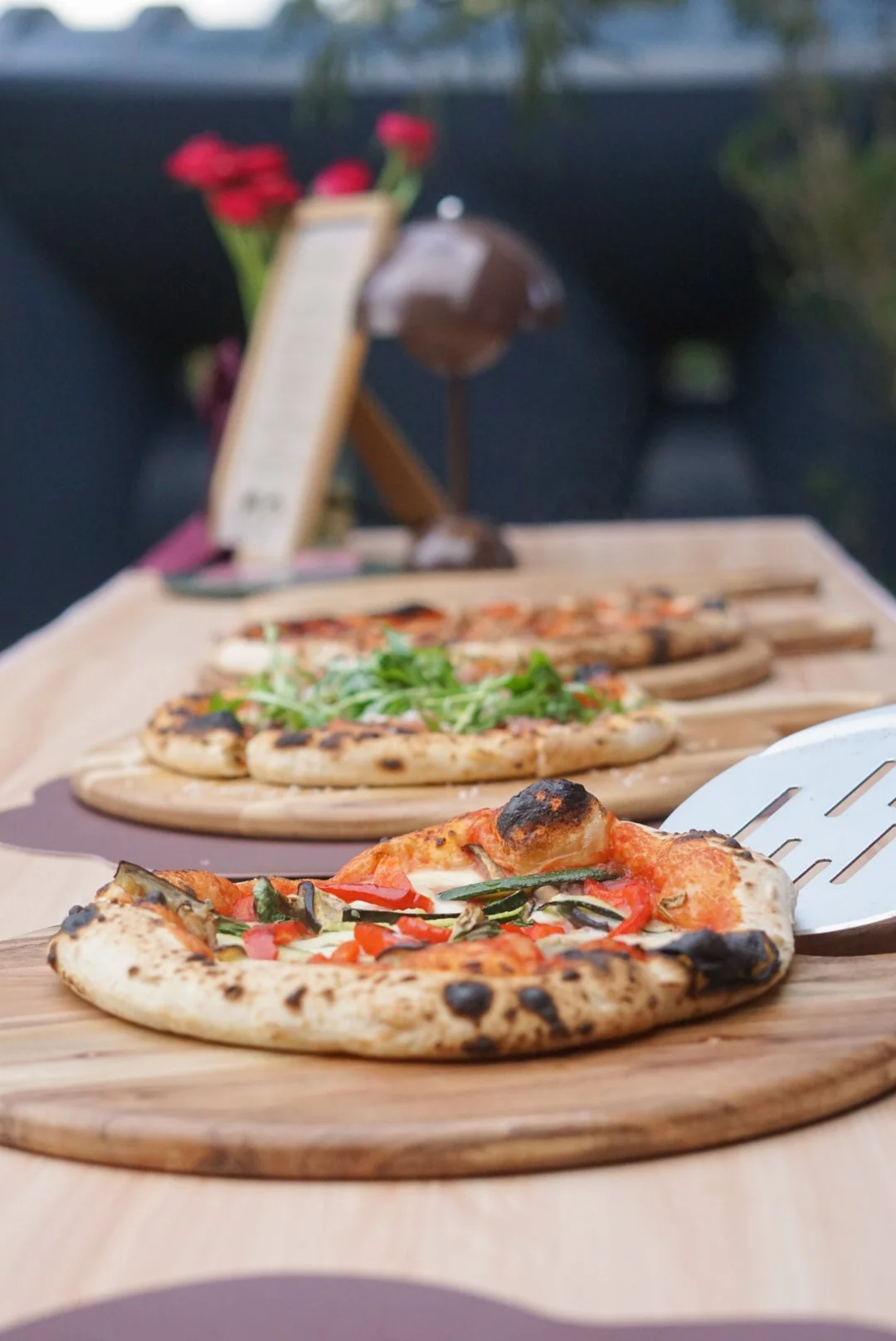 Close-up of three mini pizzas with various toppings on wooden pizza paddles, outdoors setting.