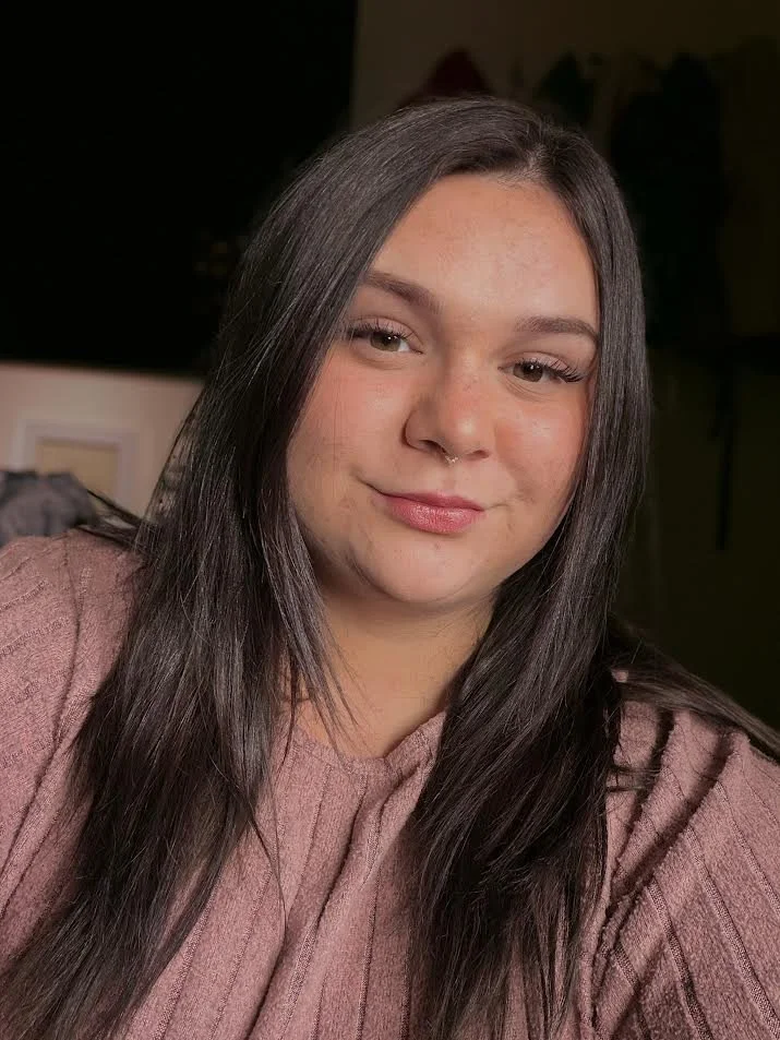 A young woman with long dark hair, wearing a mauve sweater, smiling softly at the camera in an indoor setting.