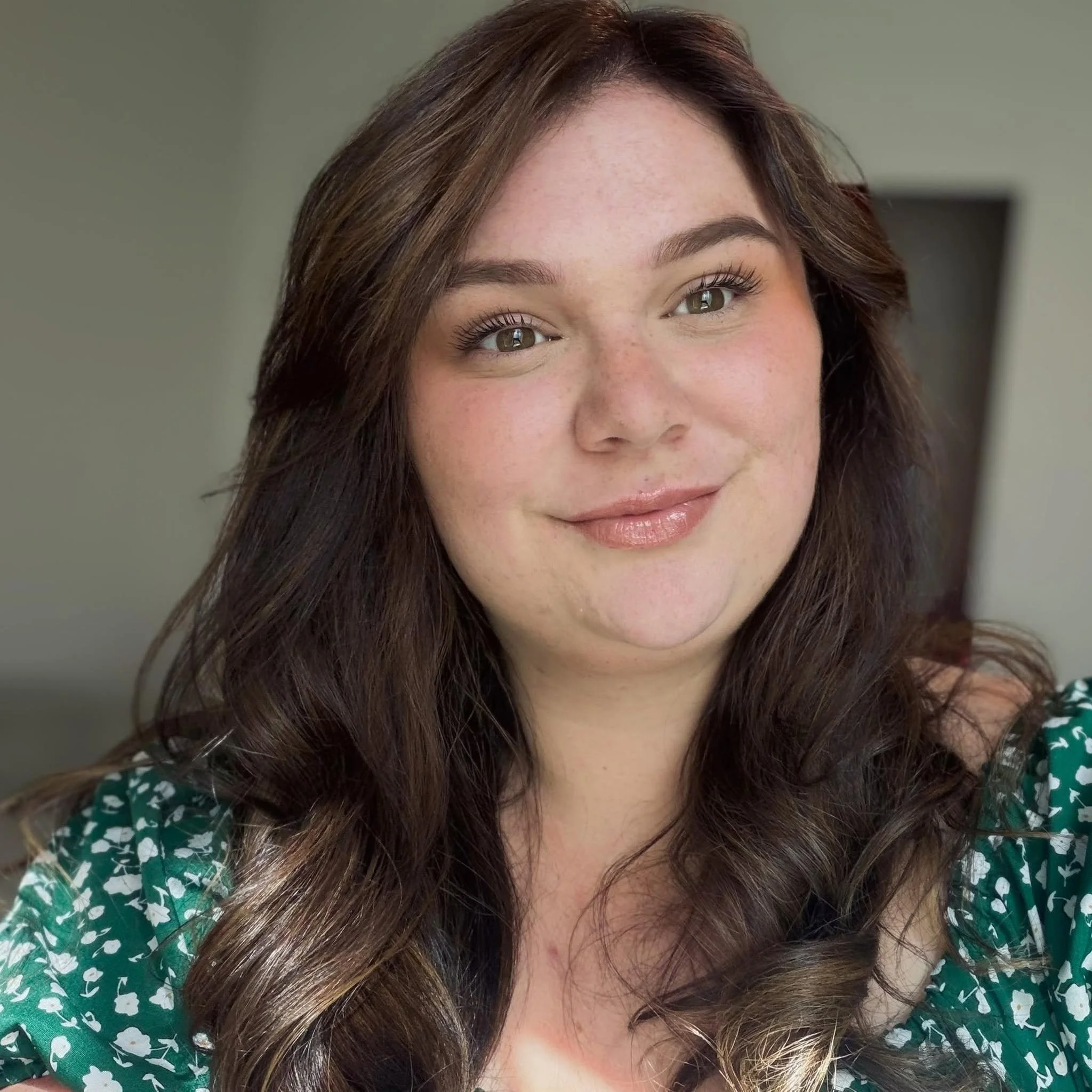 Close-up of a smiling woman with long brown hair, wearing a green top with white floral pattern.