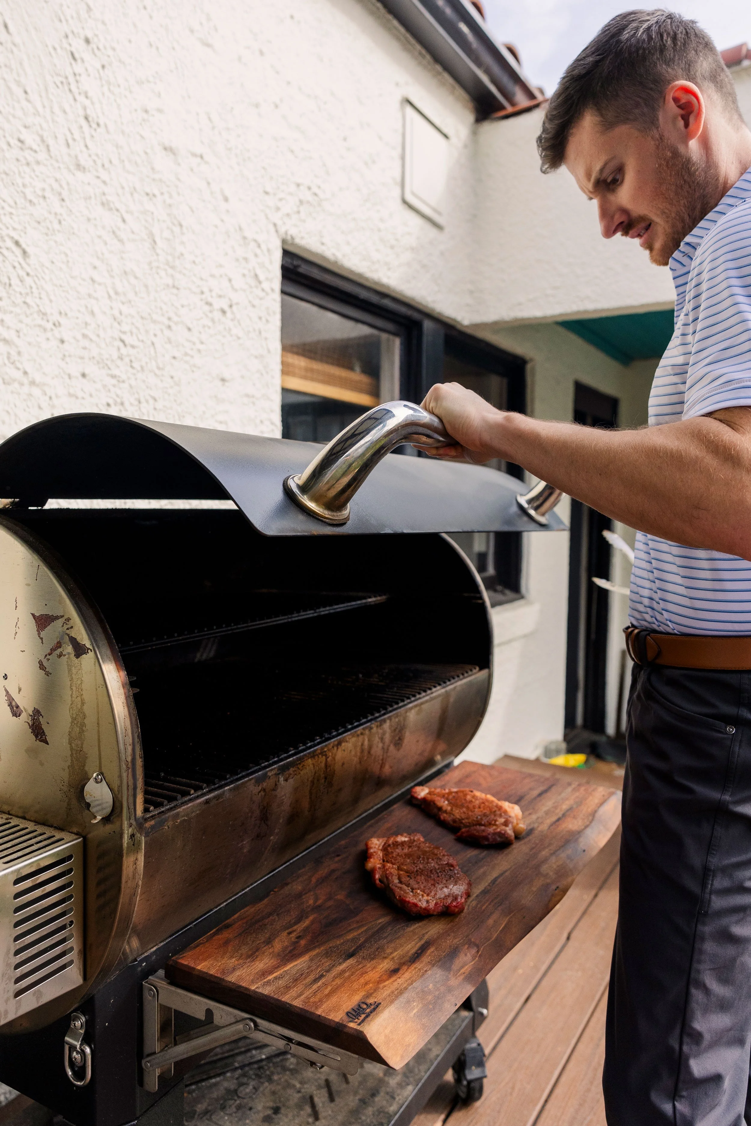 A man in a striped shirt and dark pants is grilling steaks on a barbecue grill outdoors, with some steaks on a wooden surface nearby.