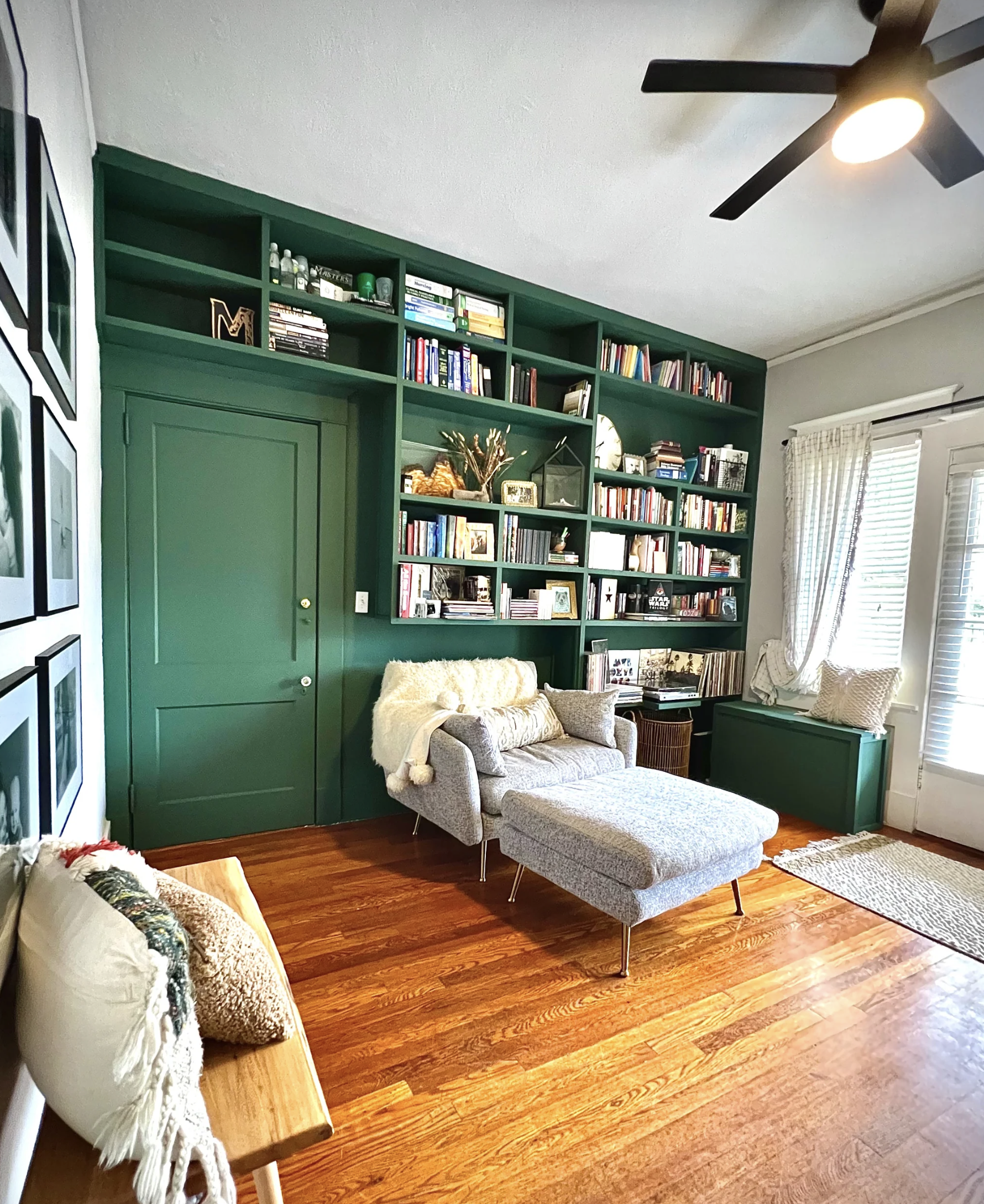 A cozy living room corner with a green wall featuring built-in bookshelves filled with books, decorations, and small plants. There is a light gray chaise lounge with a throw blanket and decorative pillow, a small side table, and a window with white b