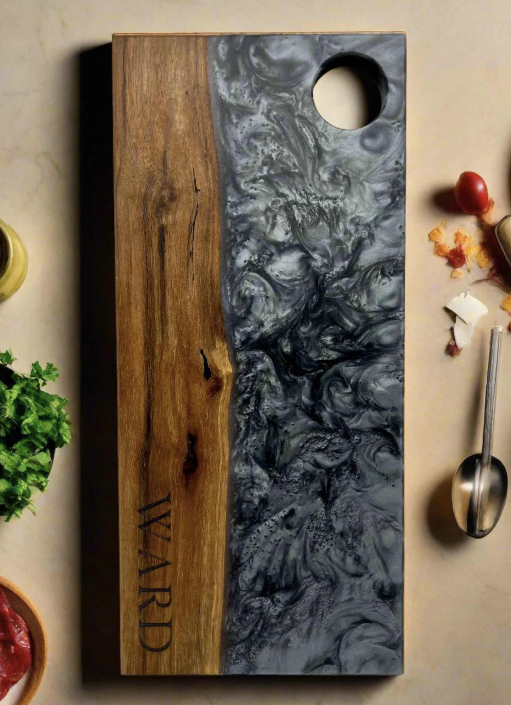 A wooden cutting board with a marble-like black and gray resin inlay. The resting part of the board is plain wood, and the inlay has the word "VIRTUE" engraved on it. Around the board are some food items and herbs, including a basil plant on the left