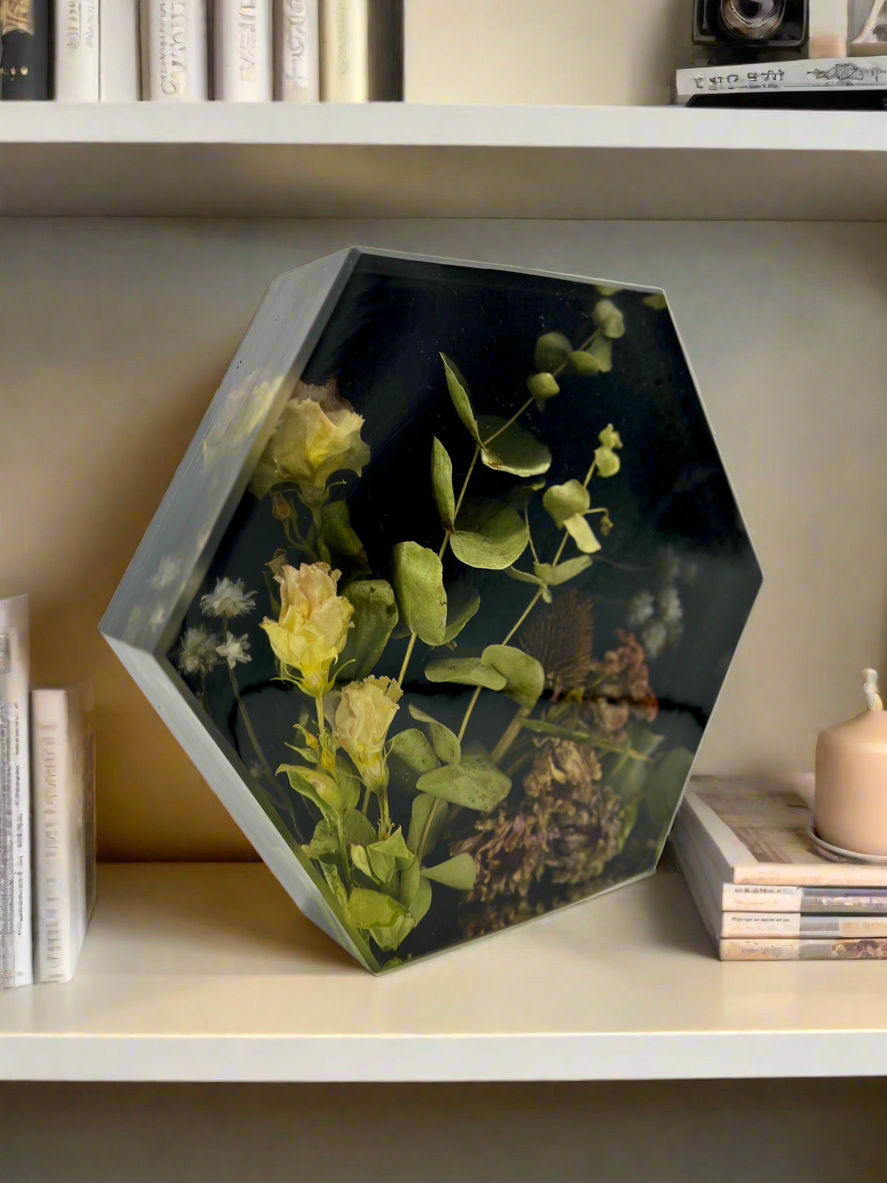 Decorative hexagonal glass box with pressed flowers and green leaves inside, placed on a white shelf next to books and a small candle.