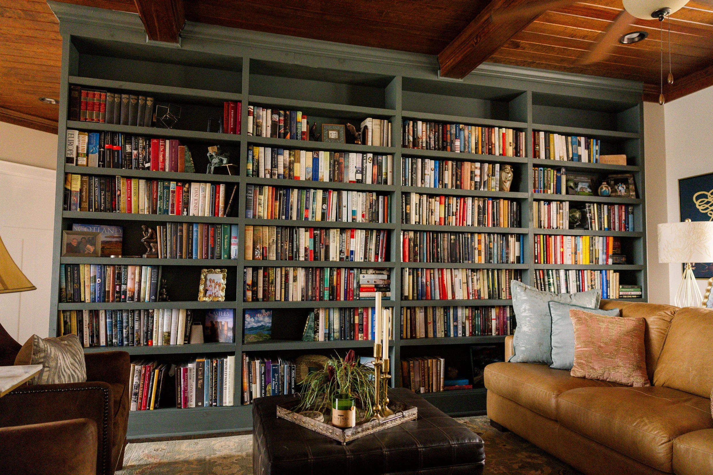 Living room with a large dark green bookshelf filled with books, decorated with picture frames and ornaments. Leather sofa with cushions, a coffee table with plants and candles, and a wooden ceiling with a ceiling fan.
