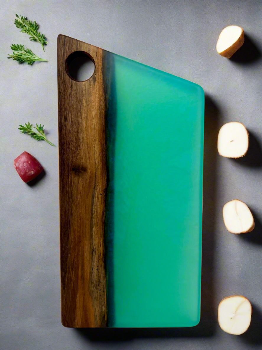 A cutting board made of wood and teal-colored resin, with herbs and vegetables around it, on a gray surface.
