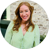 A woman with red hair smiling, wearing a light green checkered shirt, standing outside in front of a brick wall.