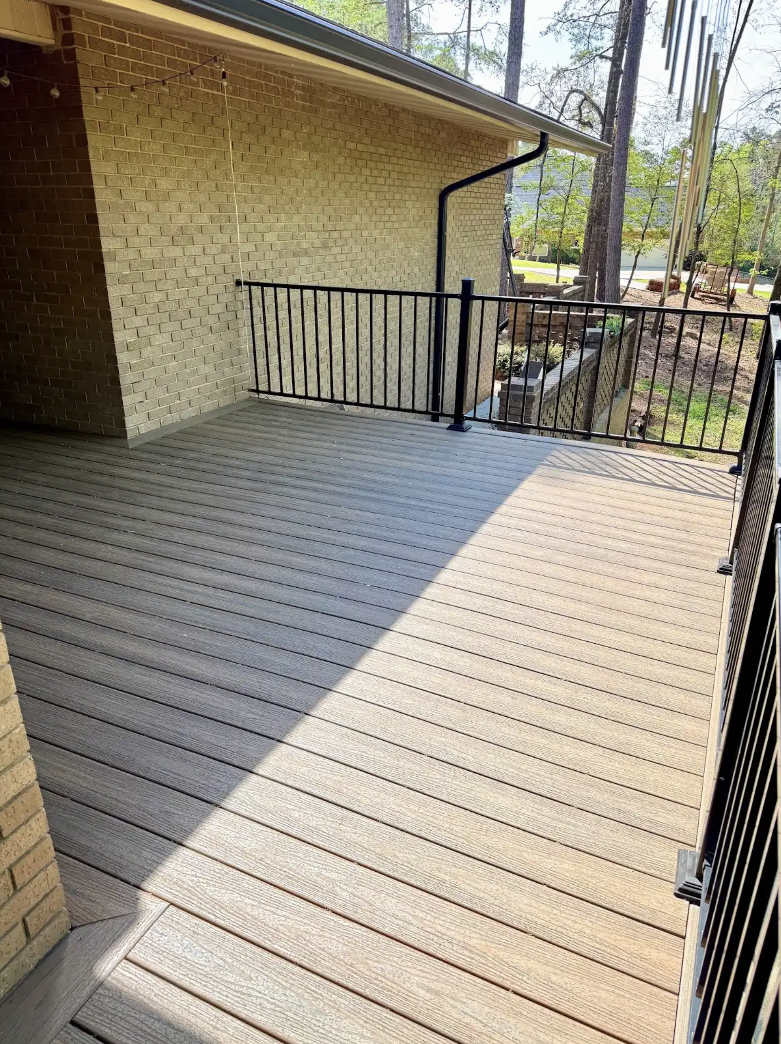 Empty wooden deck with black metal railing, attached to a house with brick exterior, overlooking a garden and trees in a backyard.