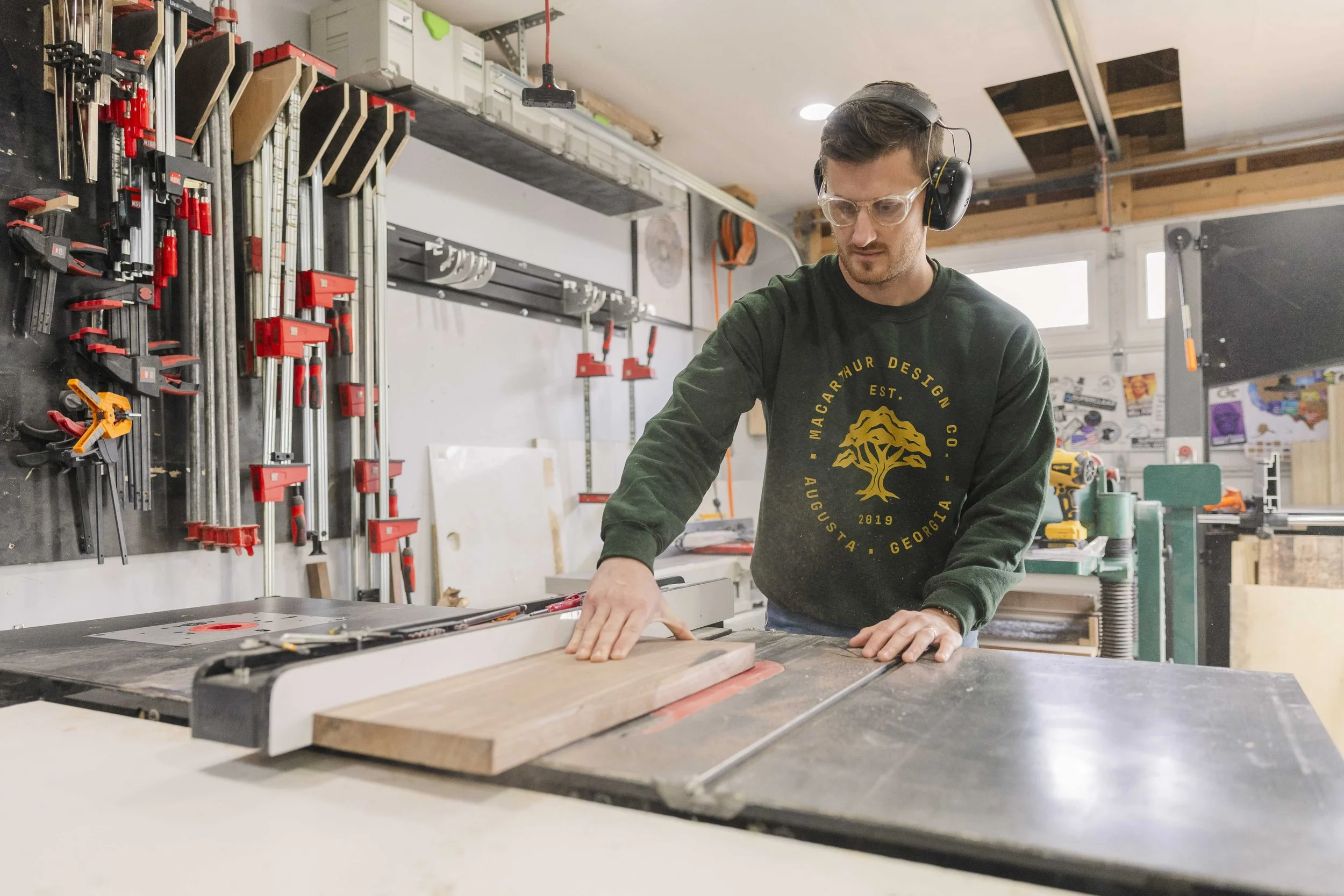 Man working in a woodworking shop, wearing safety glasses and ear protection, at a workbench with wood and tools, with clamps hanging on the wall behind him.