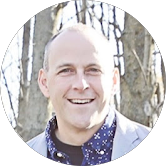 A smiling man with a bald head, wearing a light gray jacket over a patterned shirt, standing outdoors with trees in the background.