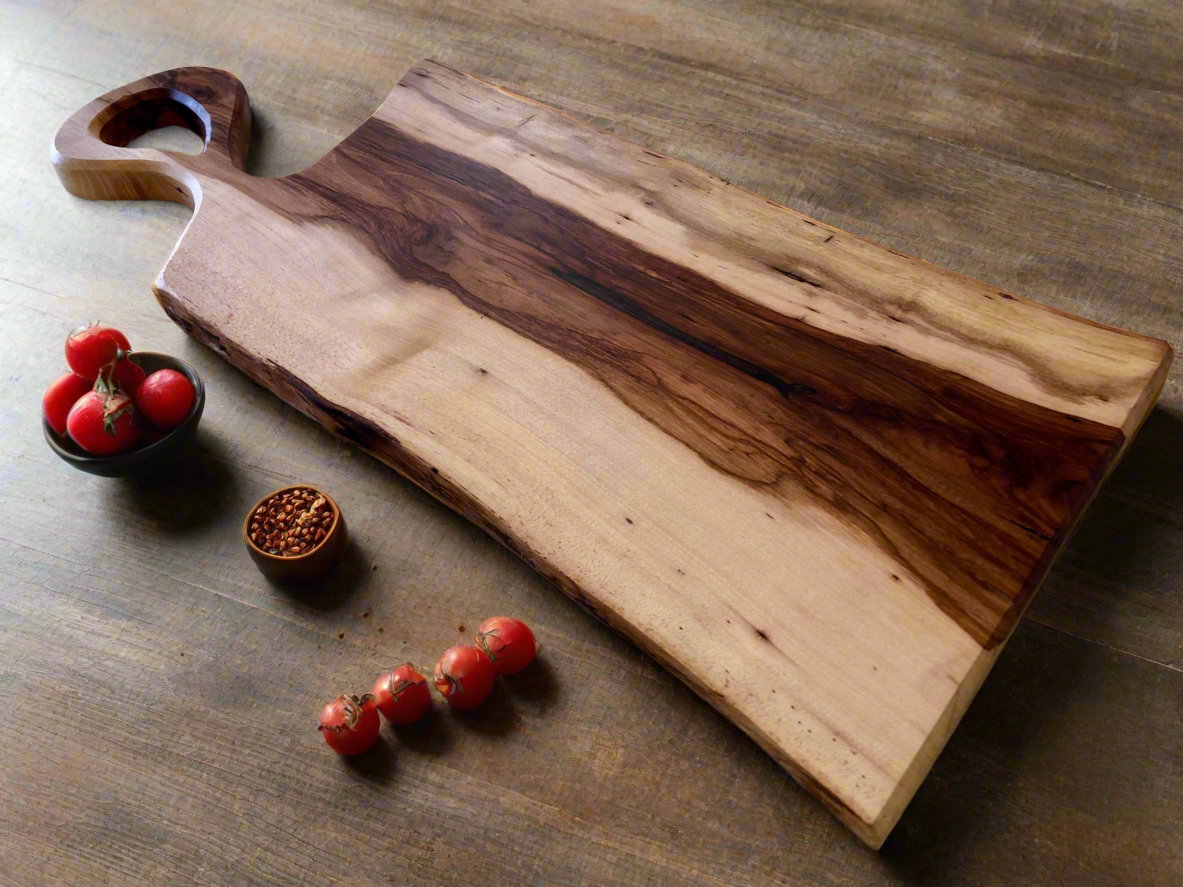 A large wooden cutting board with a handle, placed on a wooden surface, accompanied by cherry tomatoes, a small bowl of seeds, and a few more cherry tomatoes arranged around the board.