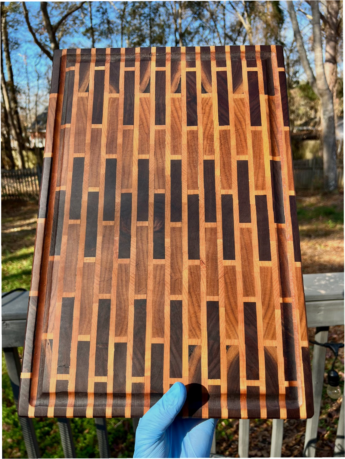 A person wearing a blue glove is holding a rectangular wooden cutting board with a geometric pattern of light and dark wood in an outdoor setting with trees and a wooden bench in the background.